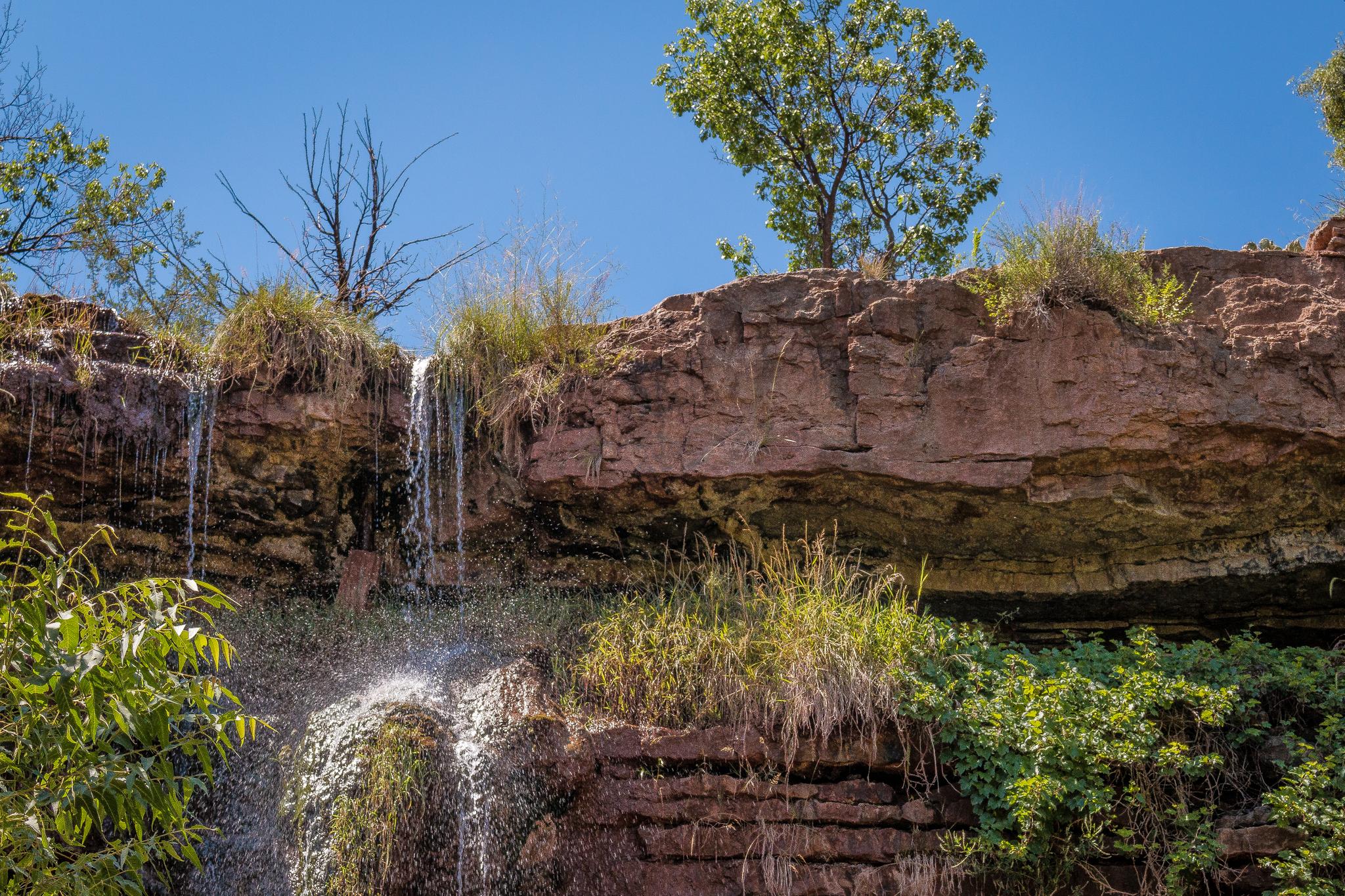 Waterfall cascades over a red rock cliff with green foliage under a blue sky.