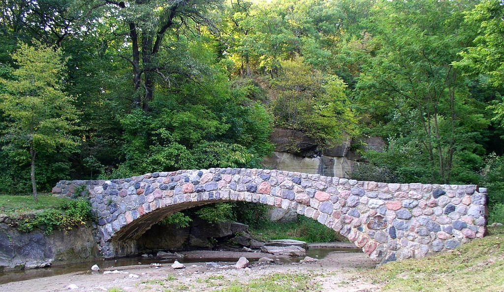 Stone arch bridge spanning a dry creek bed surrounded by green trees.