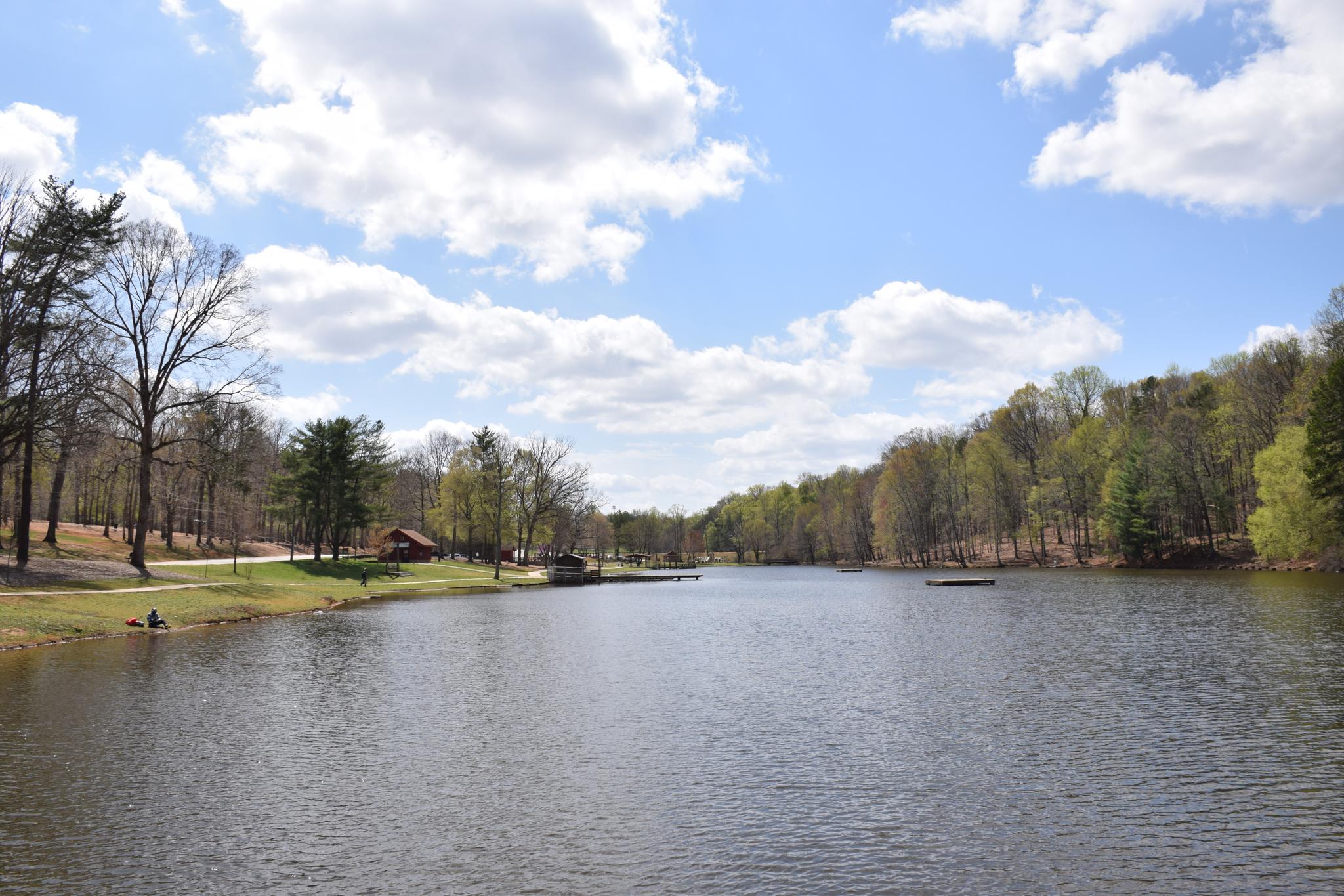 Calm lake surrounded by green trees and distant cabins under a bright, cloudy sky.