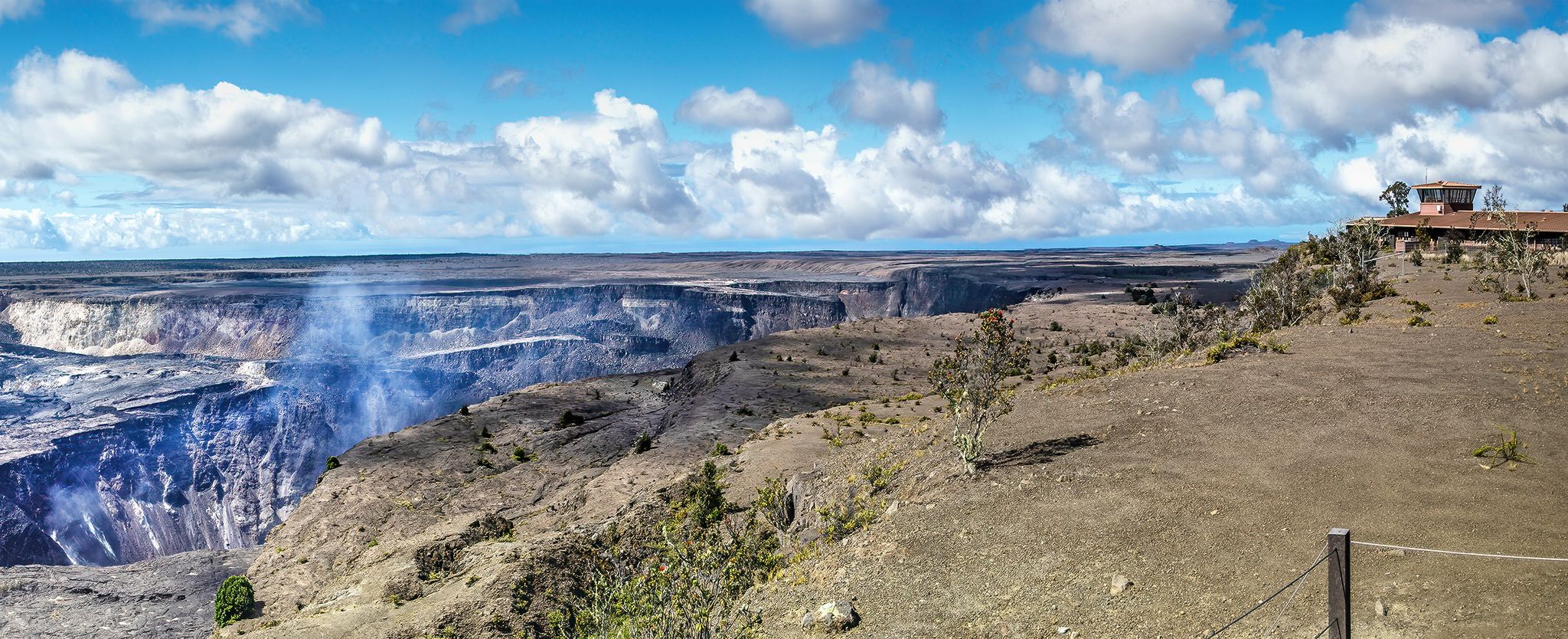 Panoramic view of a large volcanic crater with steam and a distant building. - Crater Rim Trail