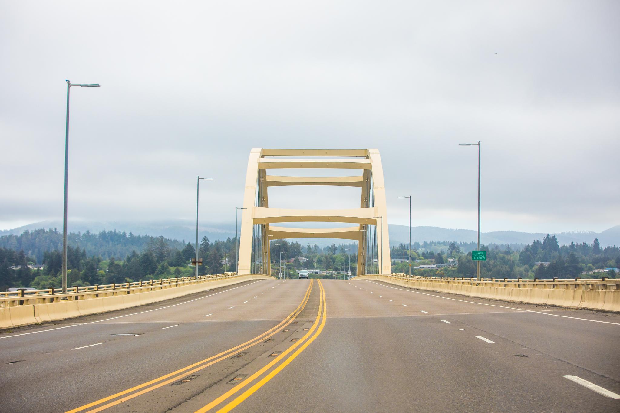 A light-colored arch bridge over a highway, with distant green hills under a cloudy sky.
