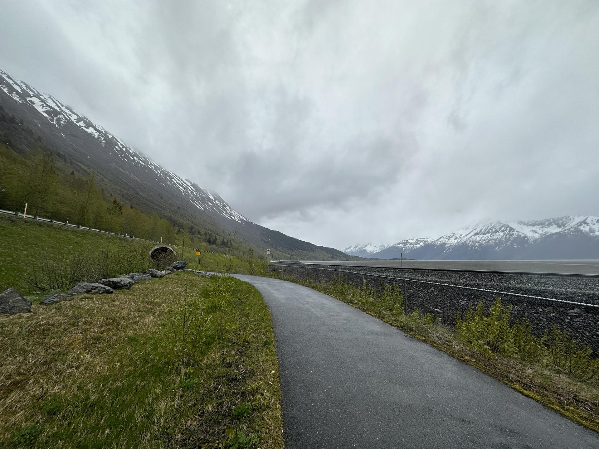 Paved path alongside a body of water with snow-capped mountains under a cloudy sky.