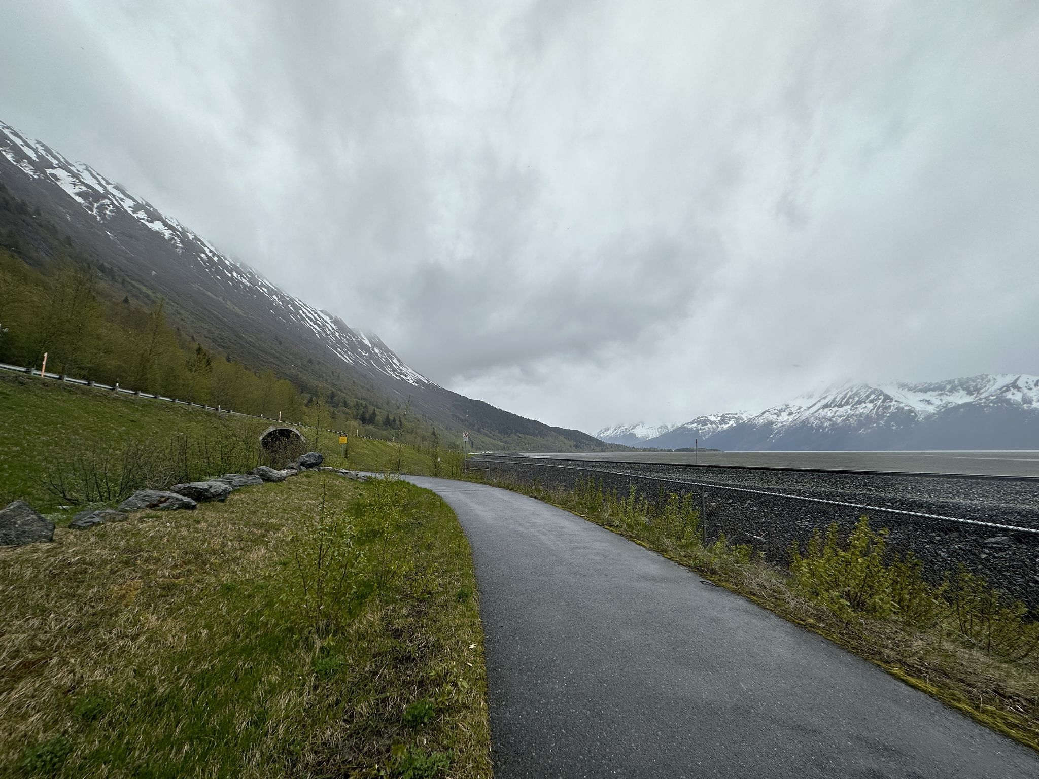 Paved path alongside a body of water with snow-capped mountains under a cloudy sky. - Bird to Gird Pathway