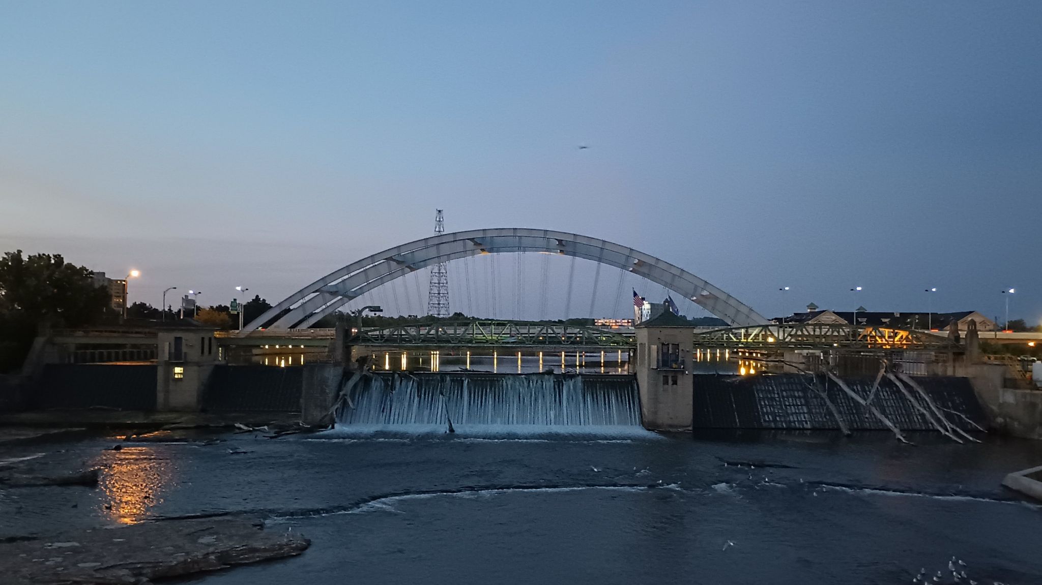 Evening view of a dam with flowing water and an illuminated arched bridge. - Genesee Riverway Trail