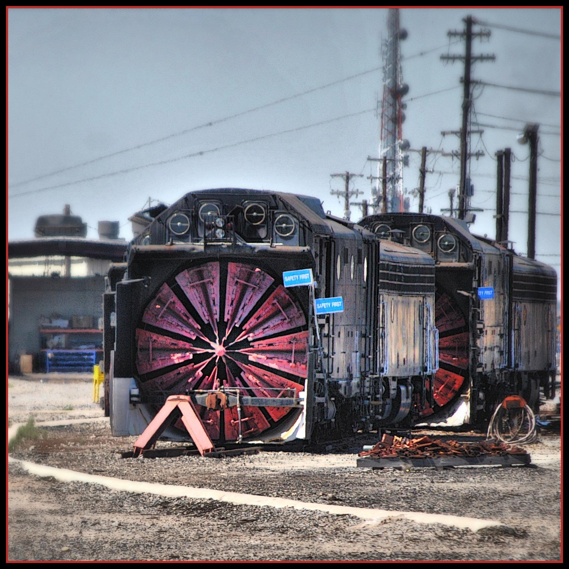 Two large, black snowplow train engines with red blades in an industrial yard.