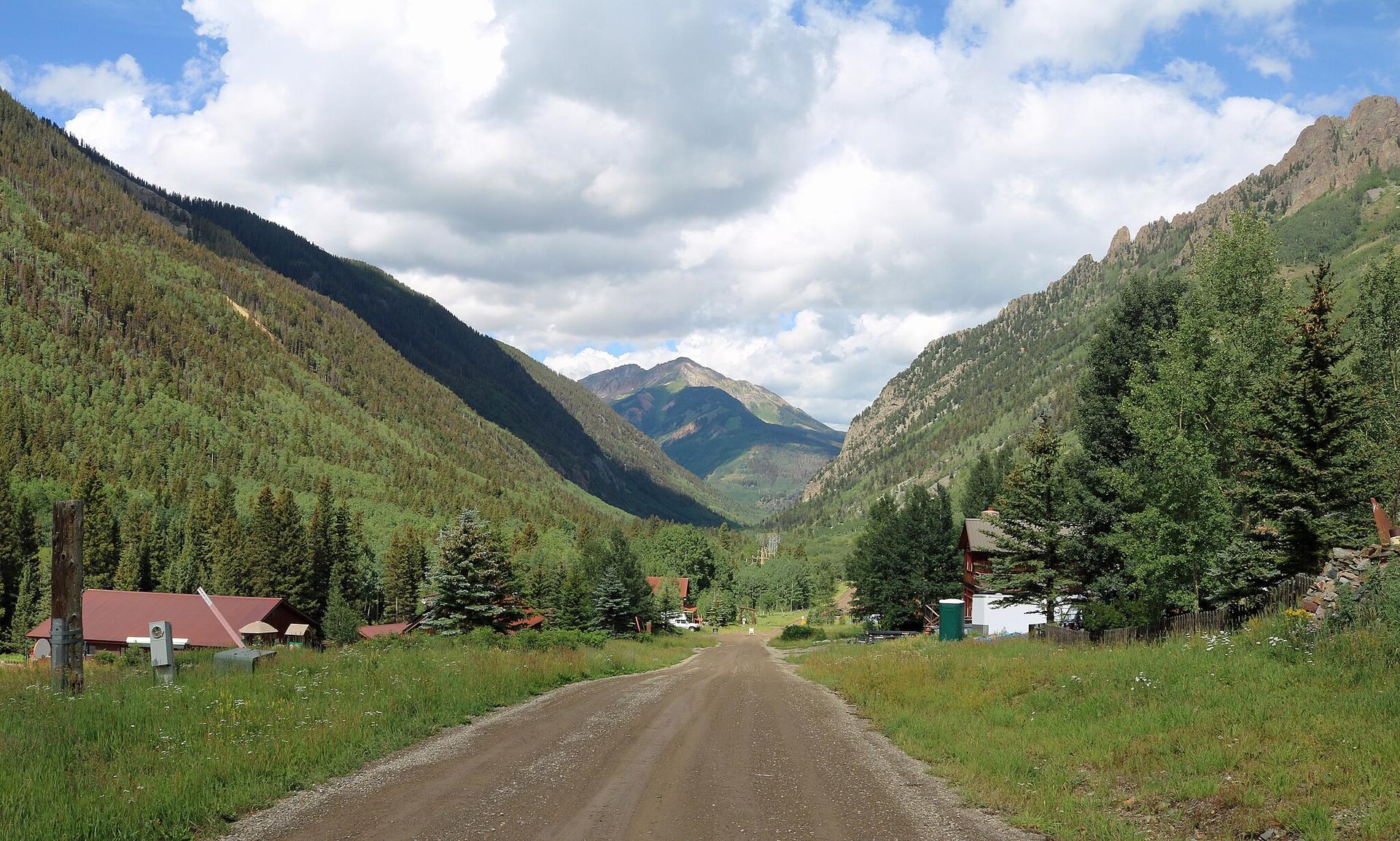 Dirt road leading through a green mountain valley with buildings under a cloudy sky.