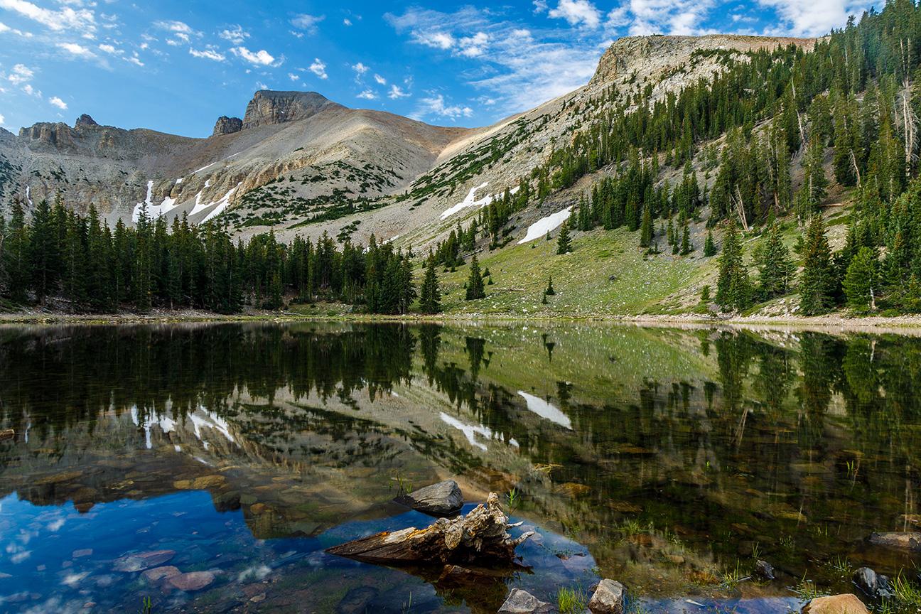 A clear mountain lake reflects forested peaks under a bright blue sky.