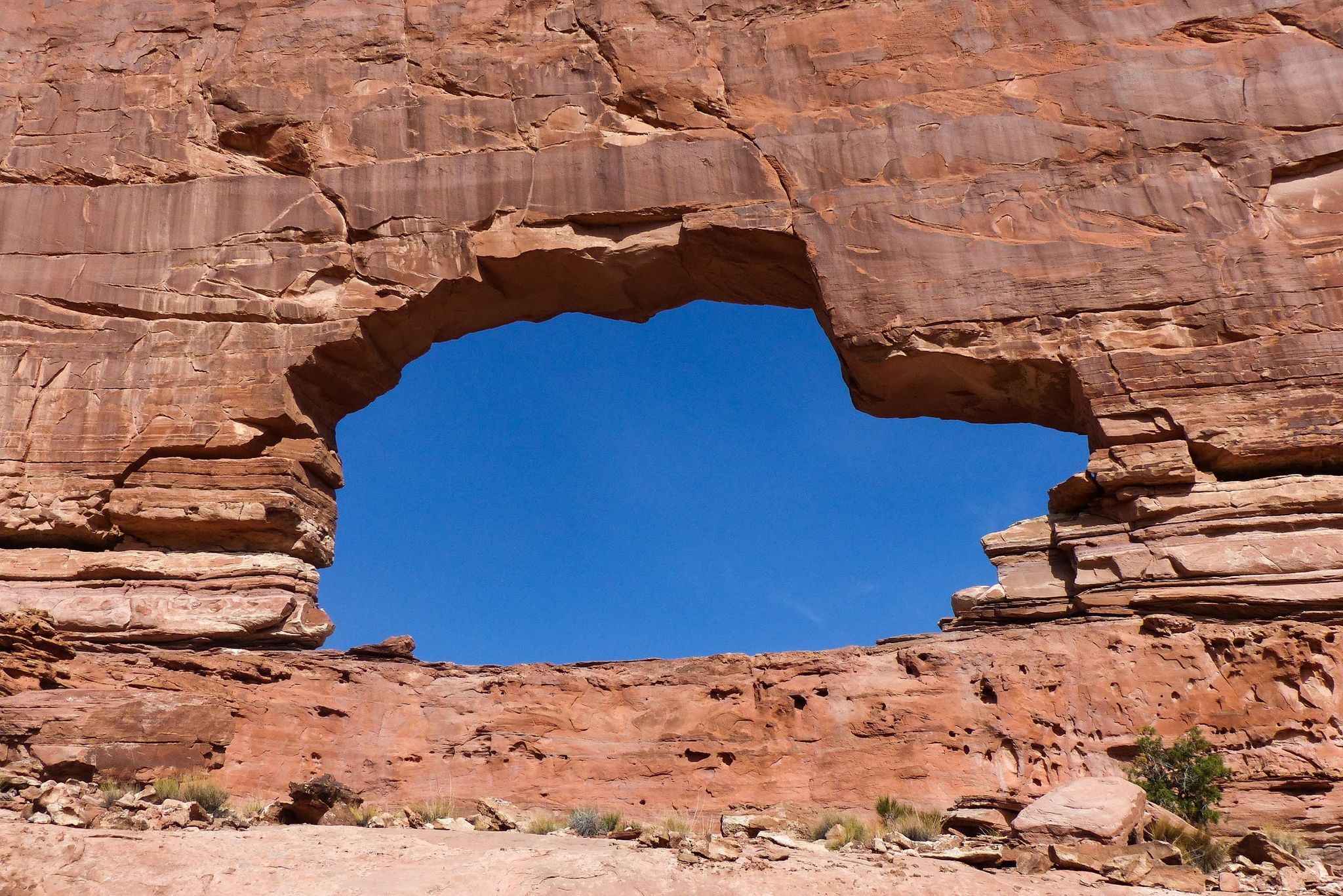 A large natural arch in red rock frames a bright blue sky. - Jeep Arch Trail