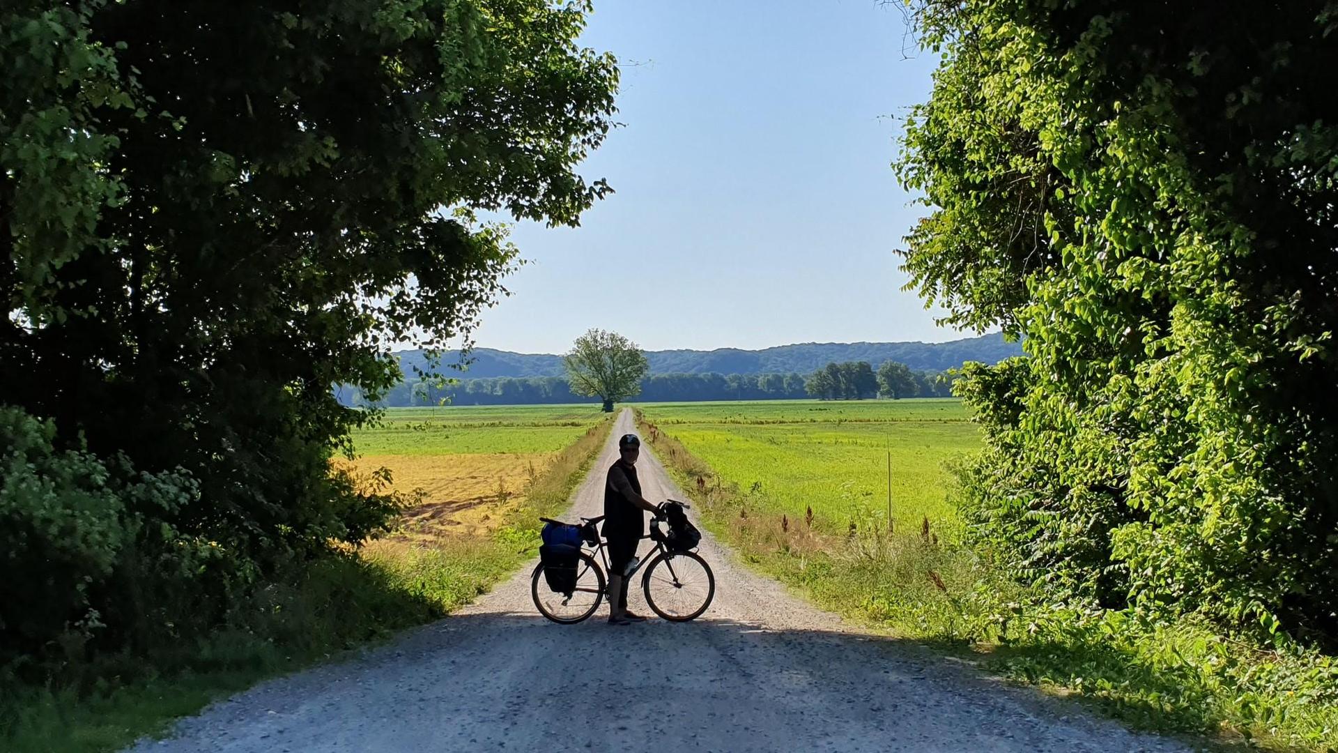 A person stands with a bicycle on a path leading to an open field.
