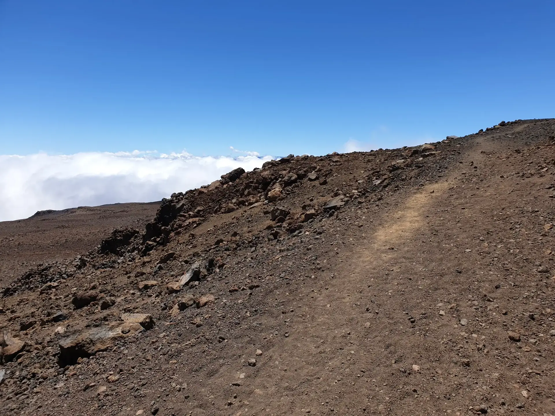 Dirt path on a rocky, brown hillside with clouds below a clear blue sky.