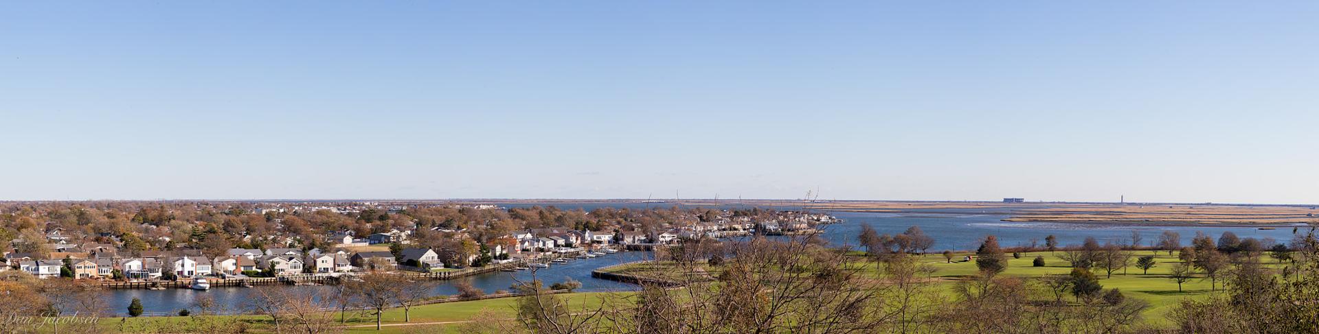 Waterfront community with houses, bare trees, and green fields under a bright blue sky.