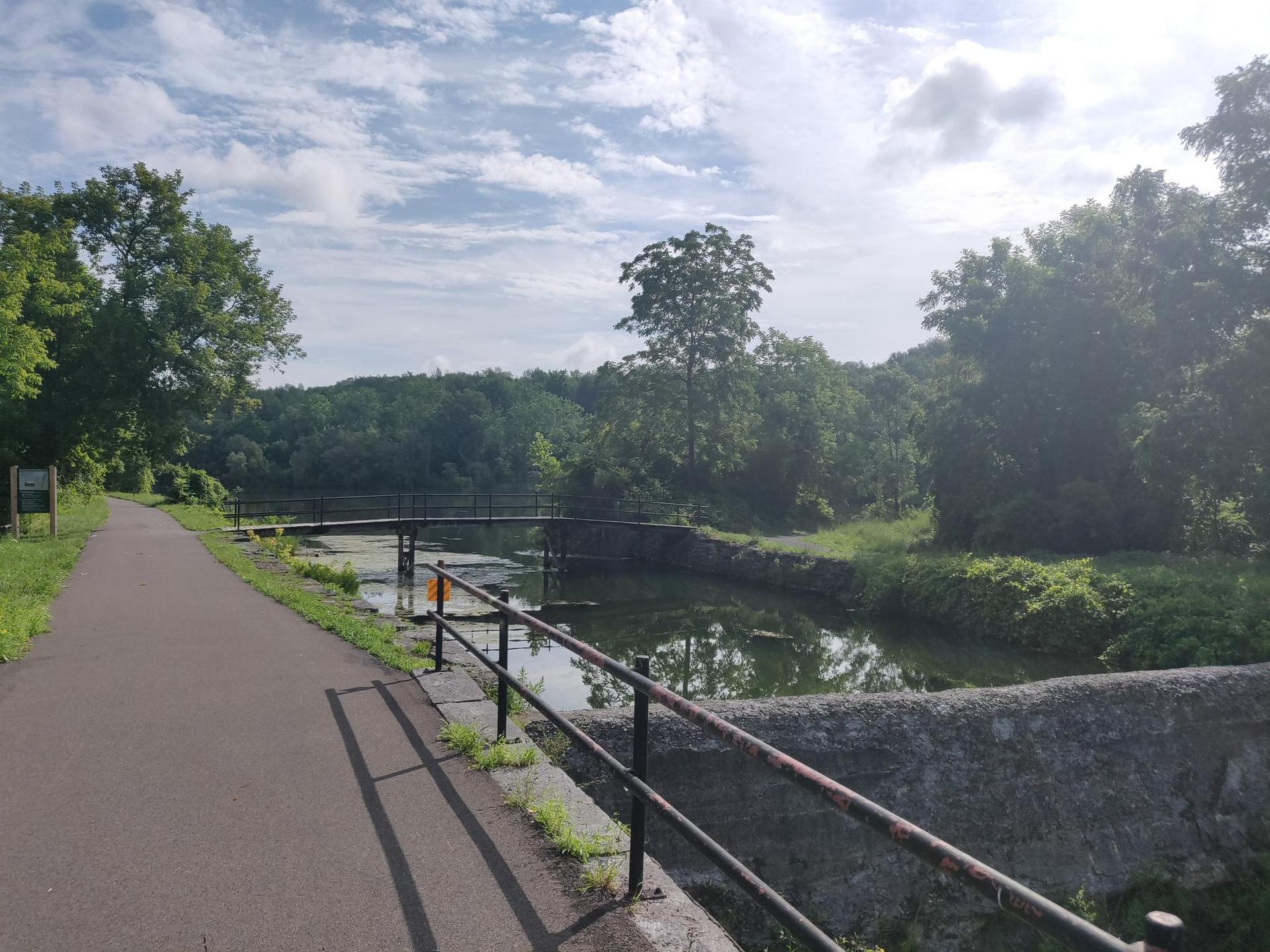 Paved path beside a canal with a small bridge, under a cloudy sky.
