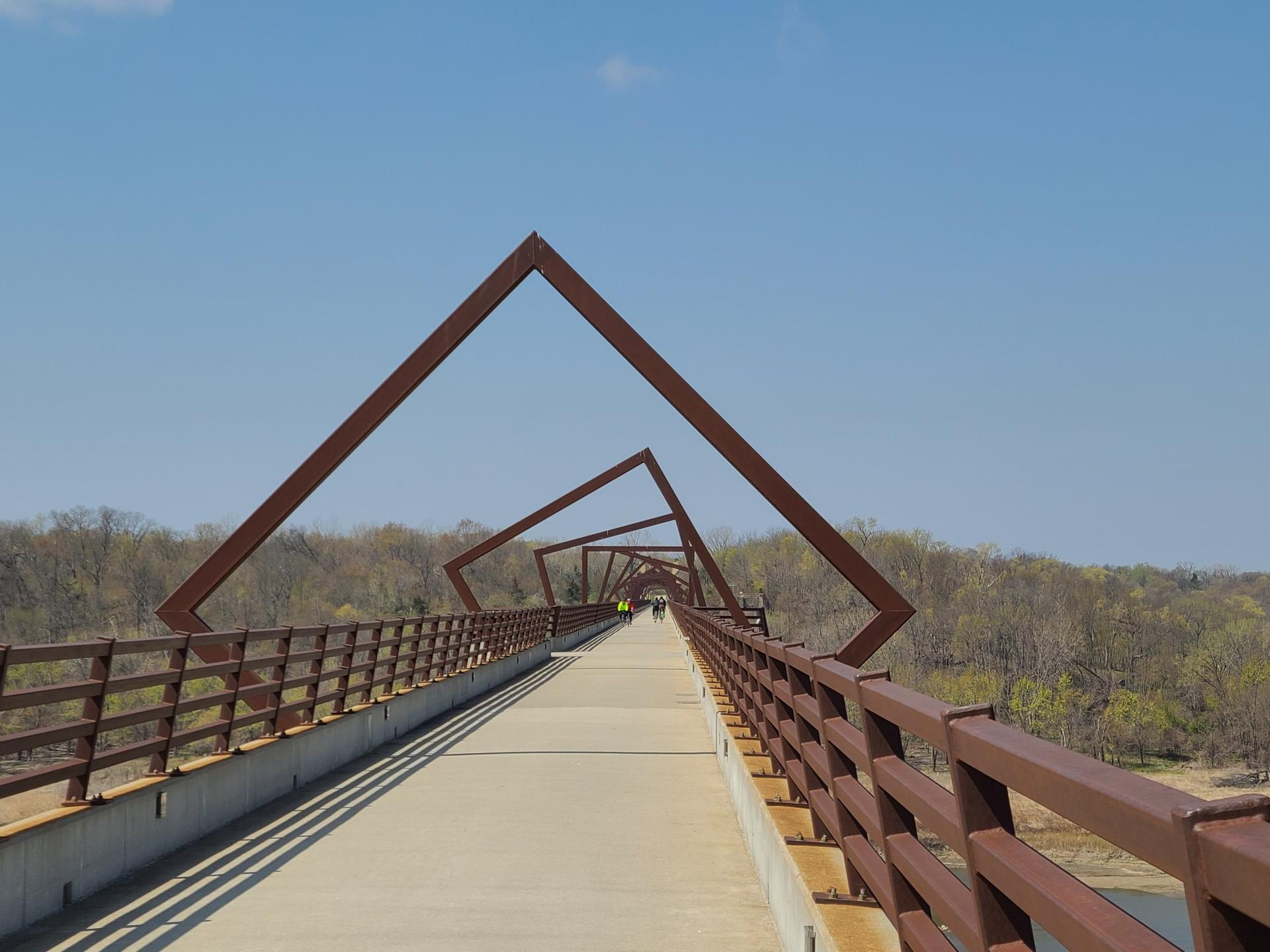 Pedestrian bridge with rusty square arches, people walking, under a blue sky.
