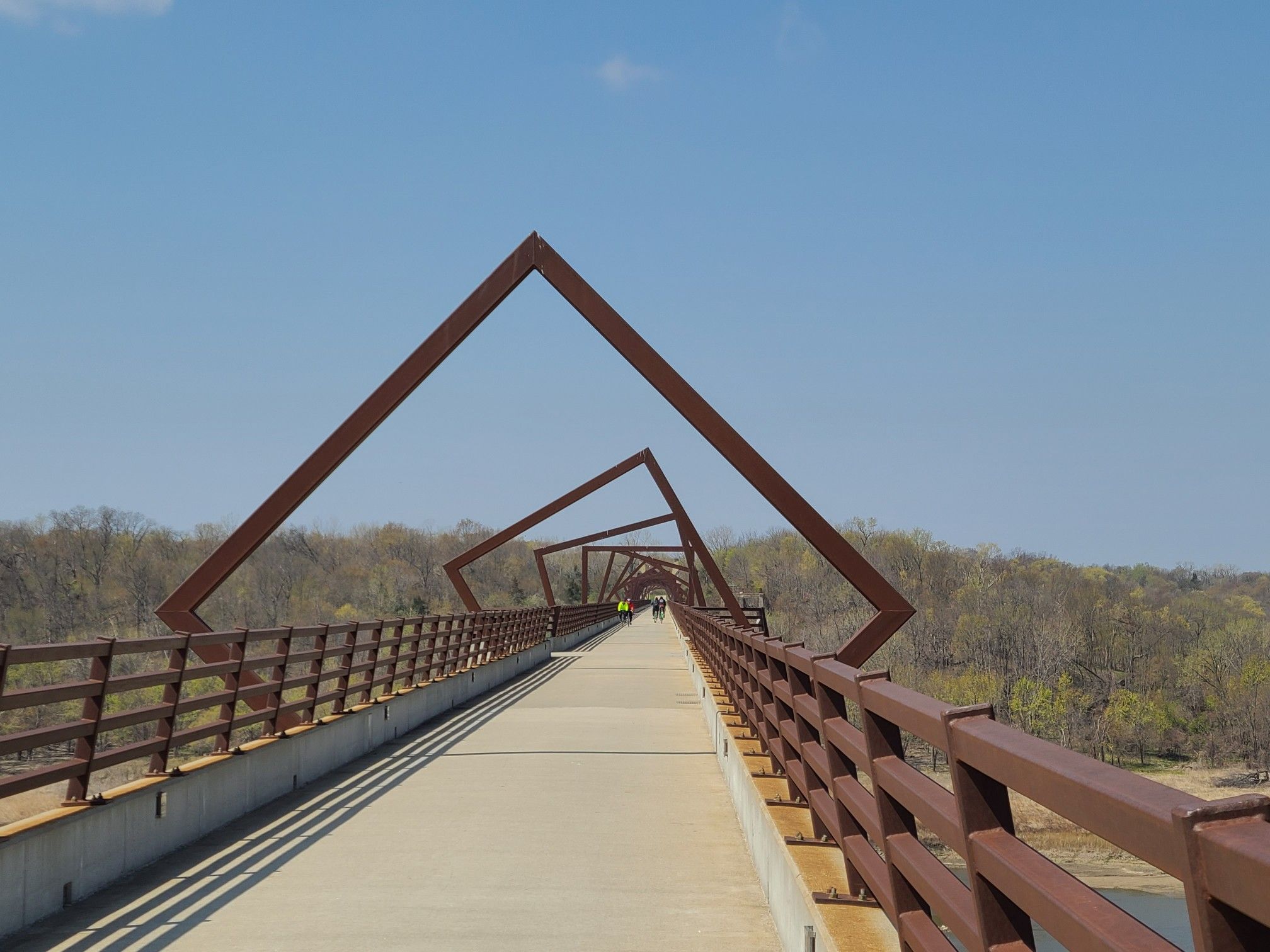 Pedestrian bridge with rusty square arches, people walking, under a blue sky. - High Trestle Trail