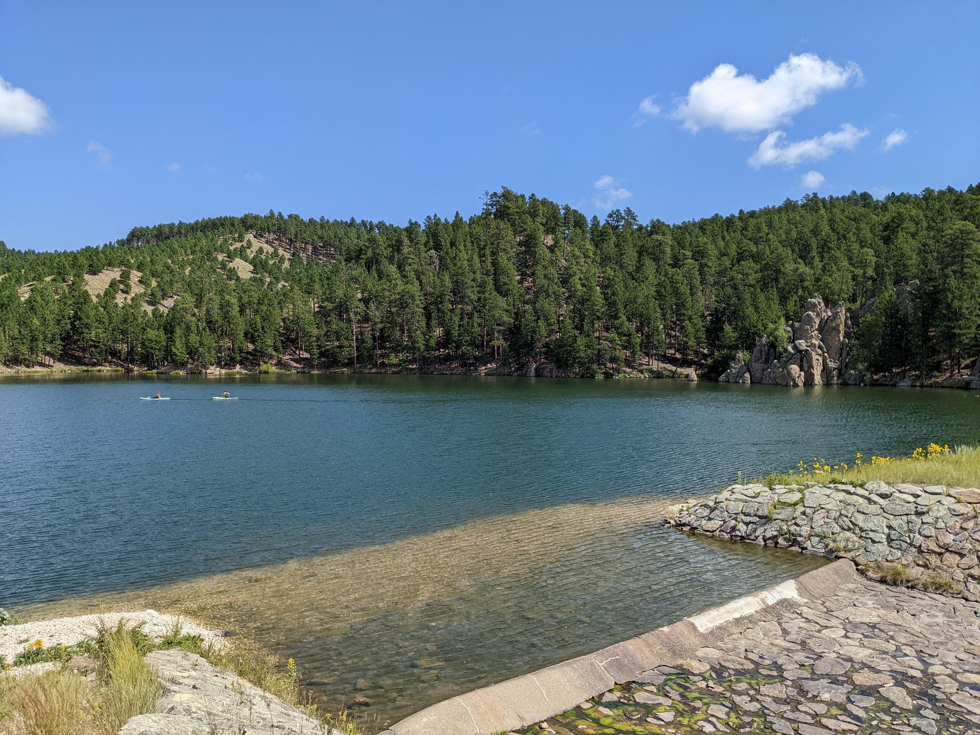 Two boats on a calm lake surrounded by forested hills under a bright blue sky.