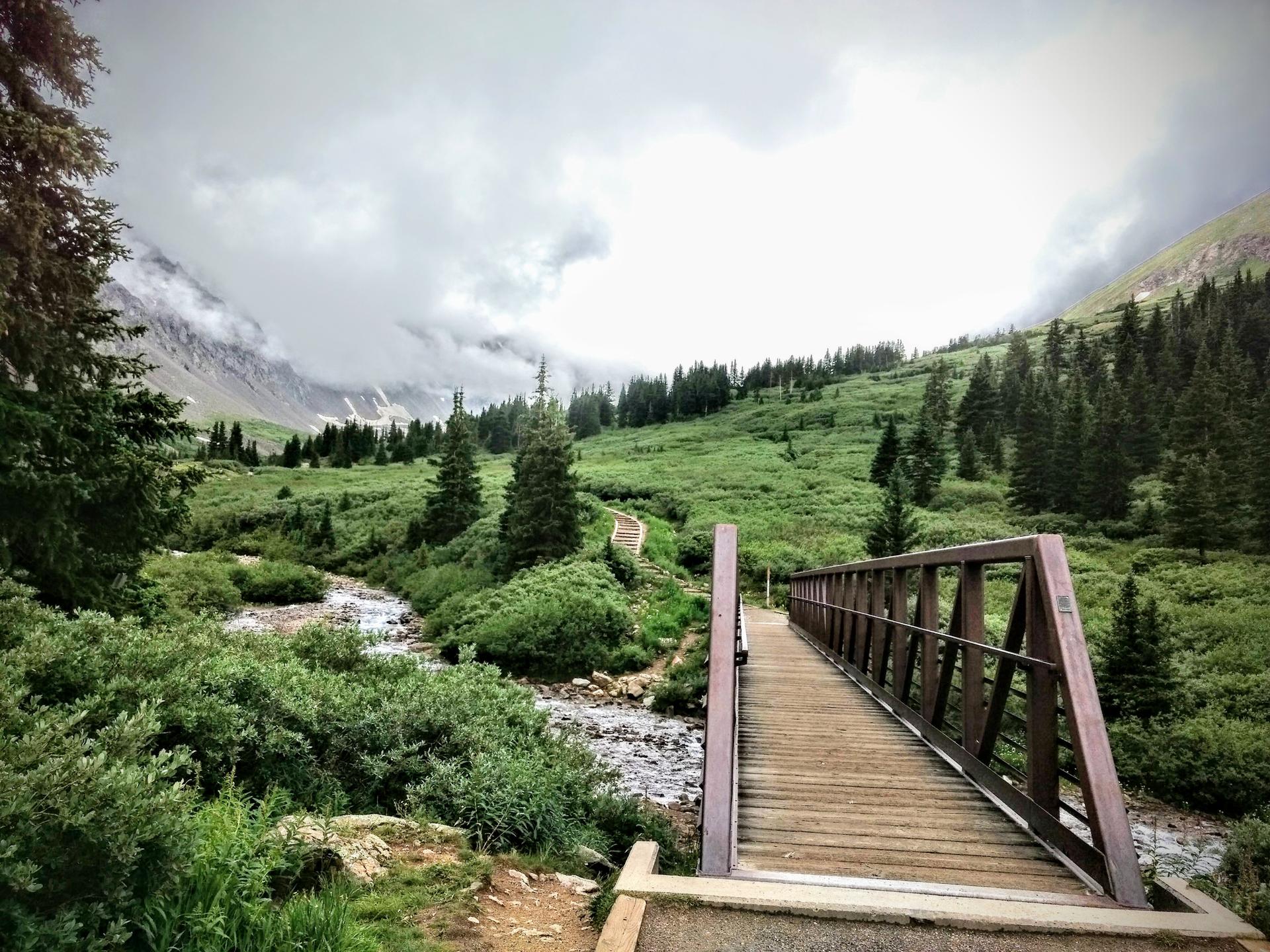 Wooden bridge over a stream in a lush green mountain valley under cloudy sky.