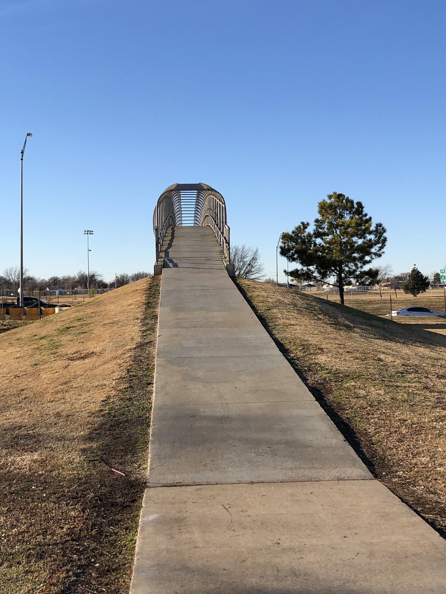 Concrete path ascending a grassy hill towards a pedestrian bridge under clear skies.