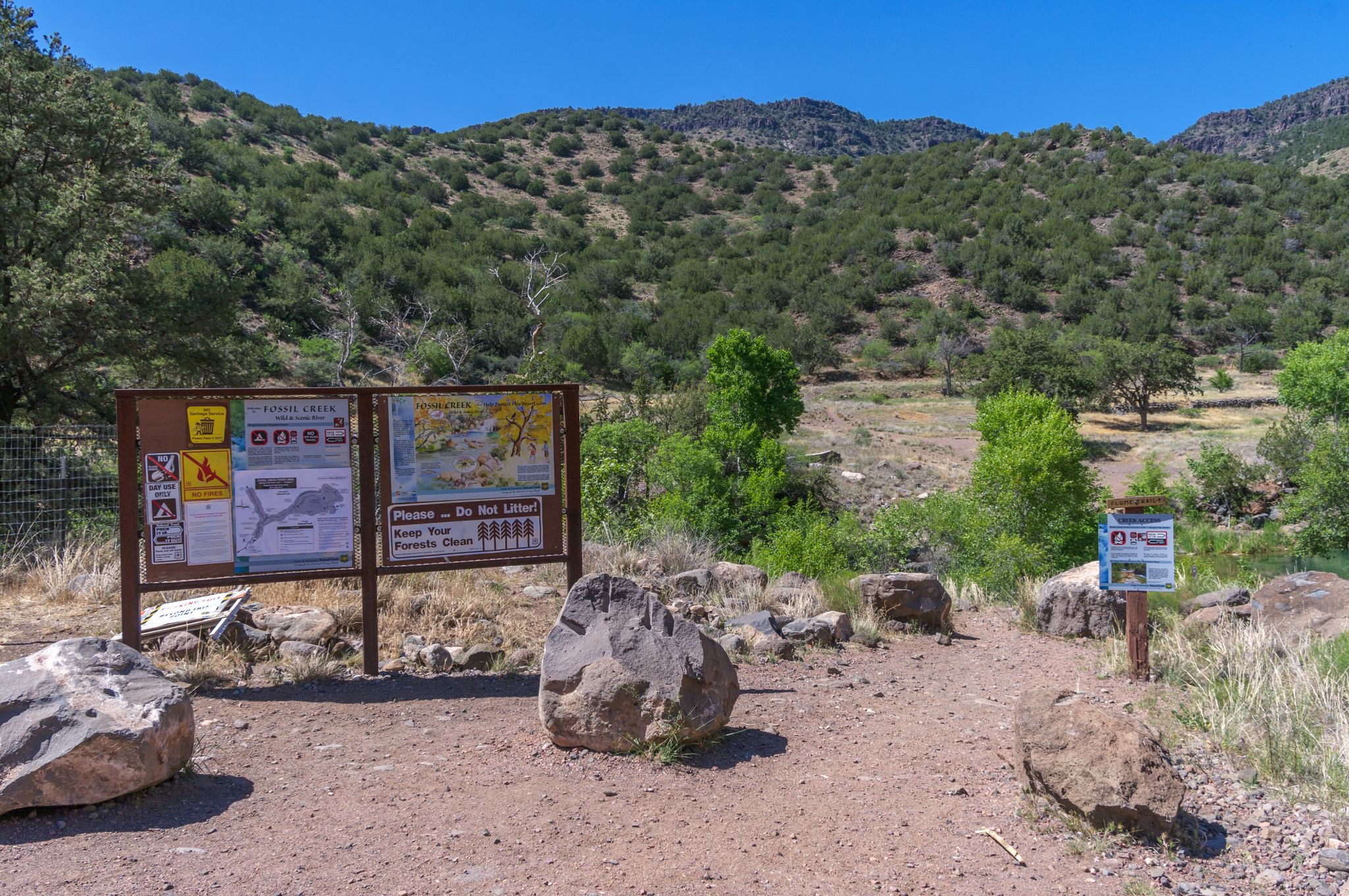 An information board and trail sign stand on a dirt path before sunny, green hills. - Flume Trail