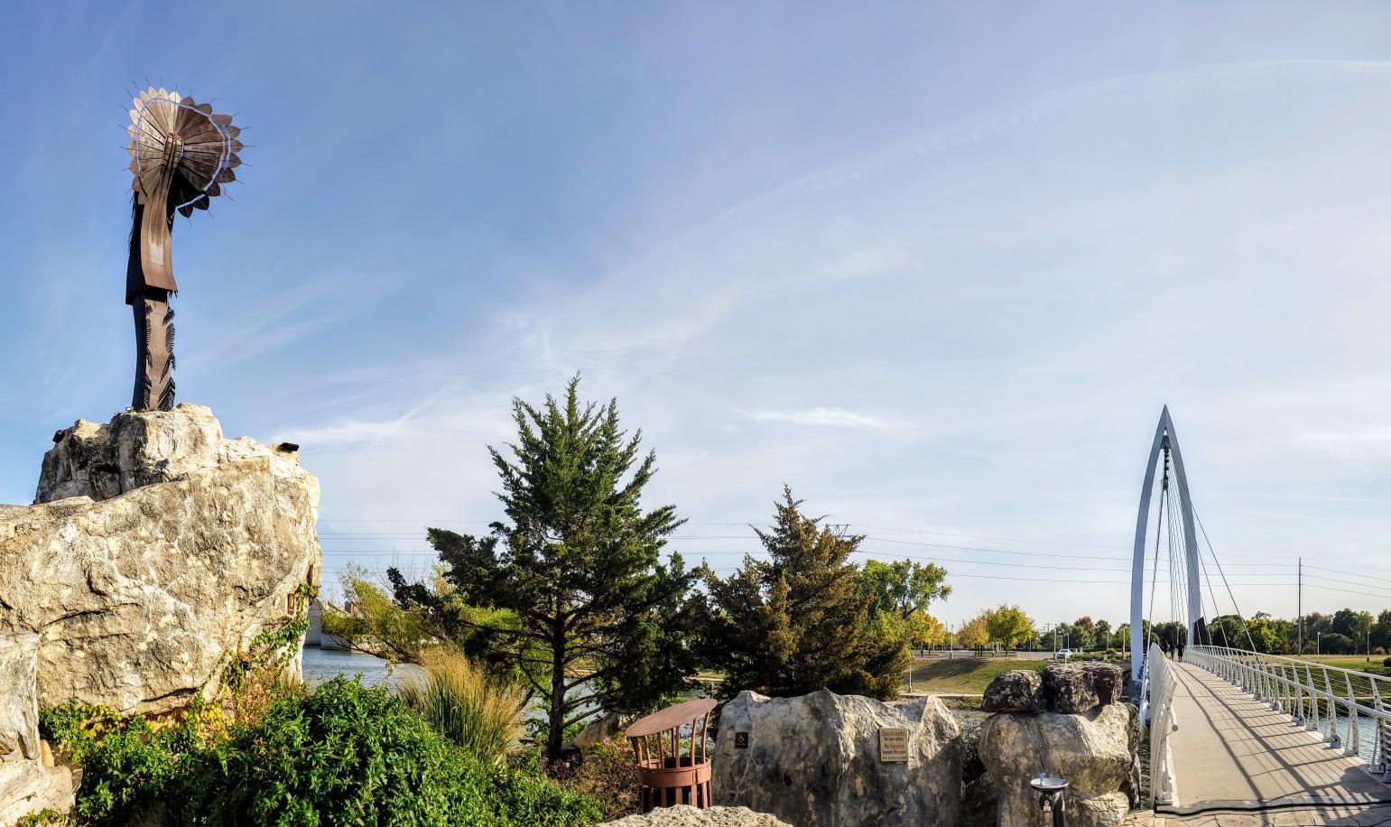Metal sculpture on rocks overlooks a river and modern pedestrian bridge under blue sky.