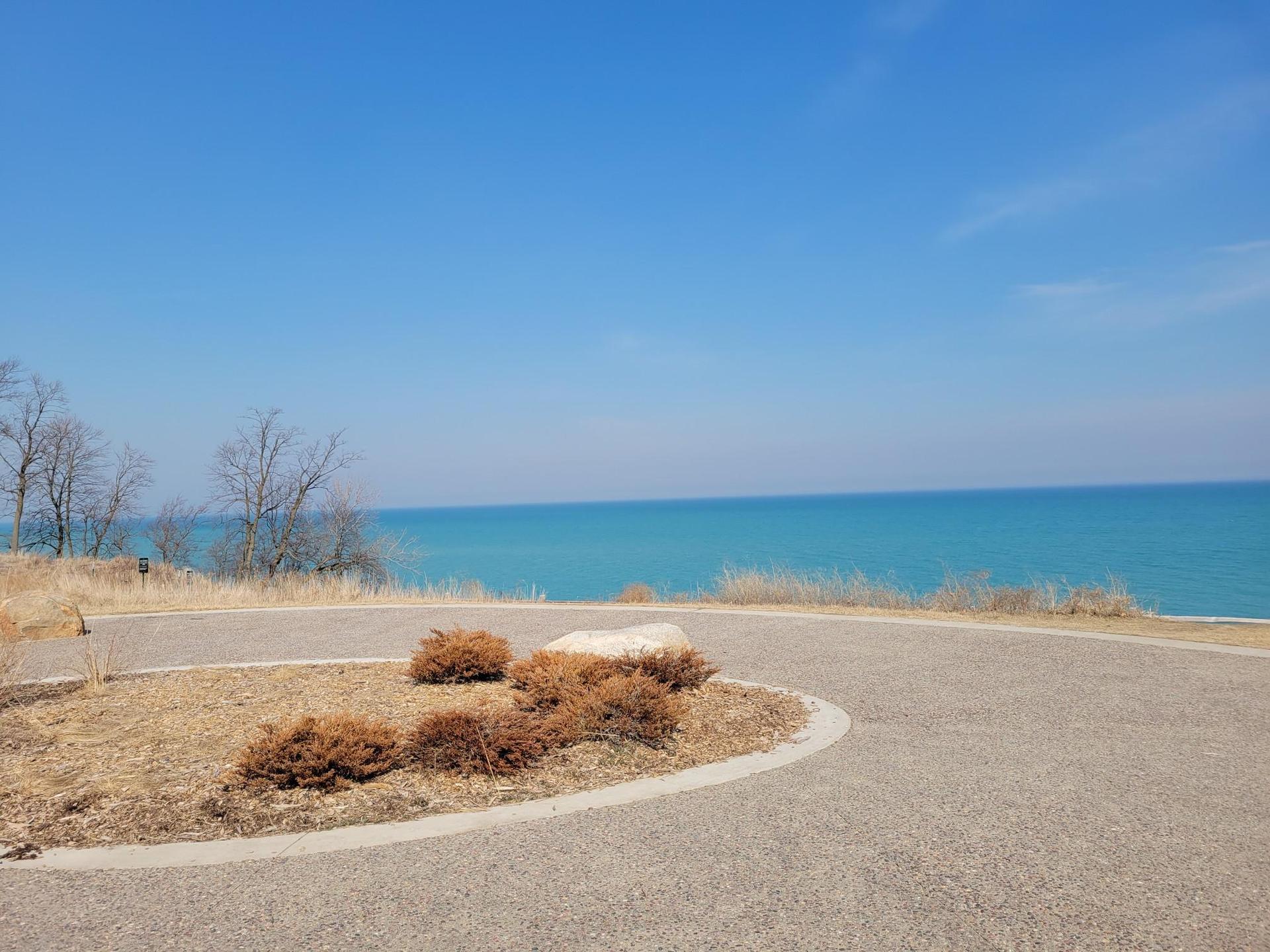 Paved overlook with dry bushes, overlooking a vast blue body of water.