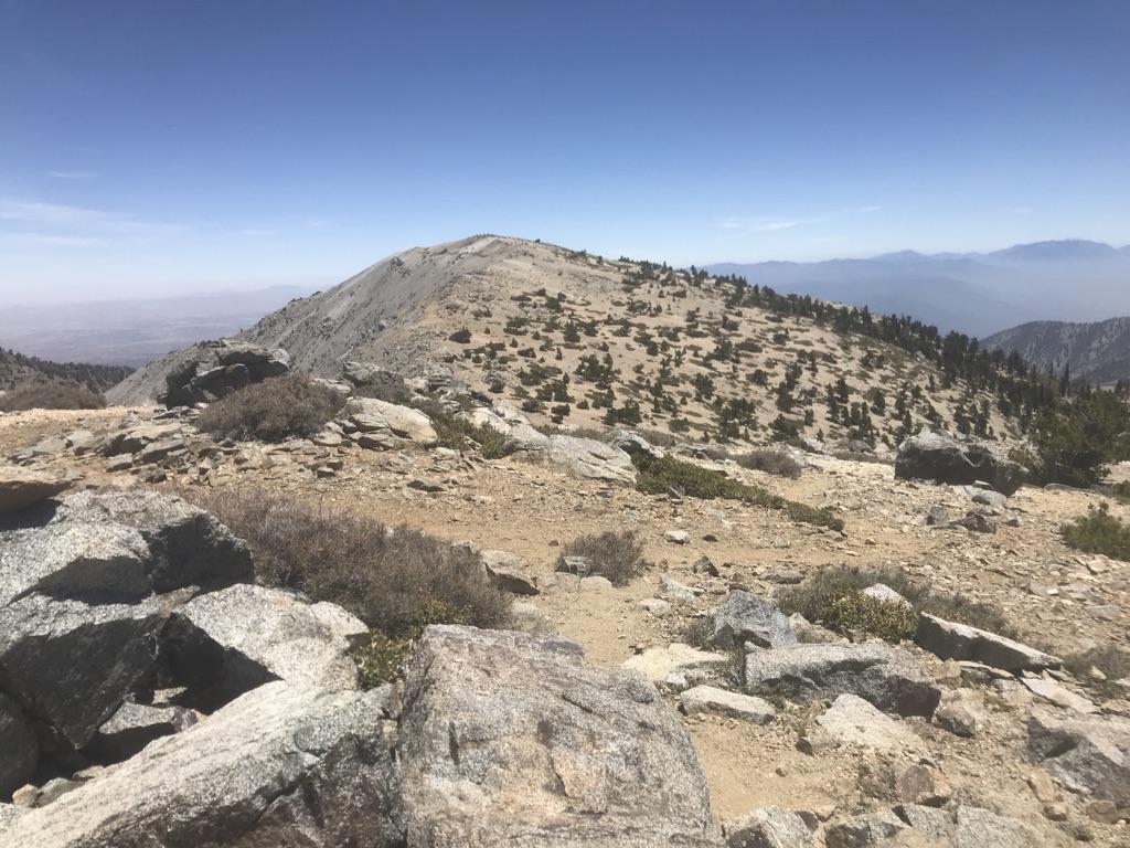 Rocky mountain peak under clear blue sky, sparse vegetation, distant hazy mountains.
