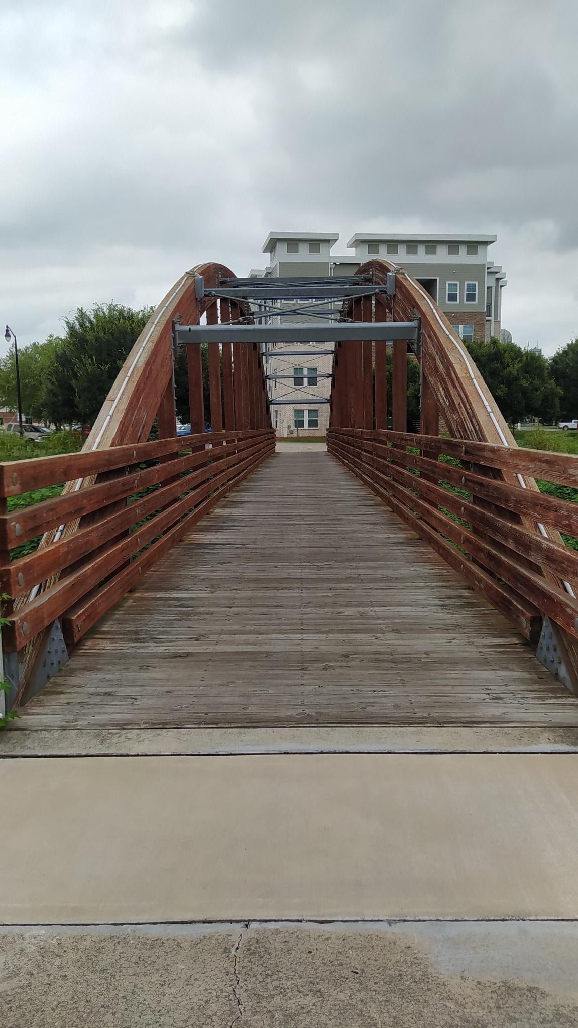 A wooden arched pedestrian bridge leads towards modern buildings under an overcast sky.