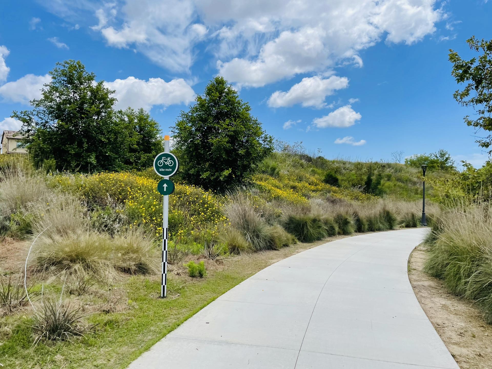 Paved path curving right, with a bike trail sign and green hills under a cloudy sky.
