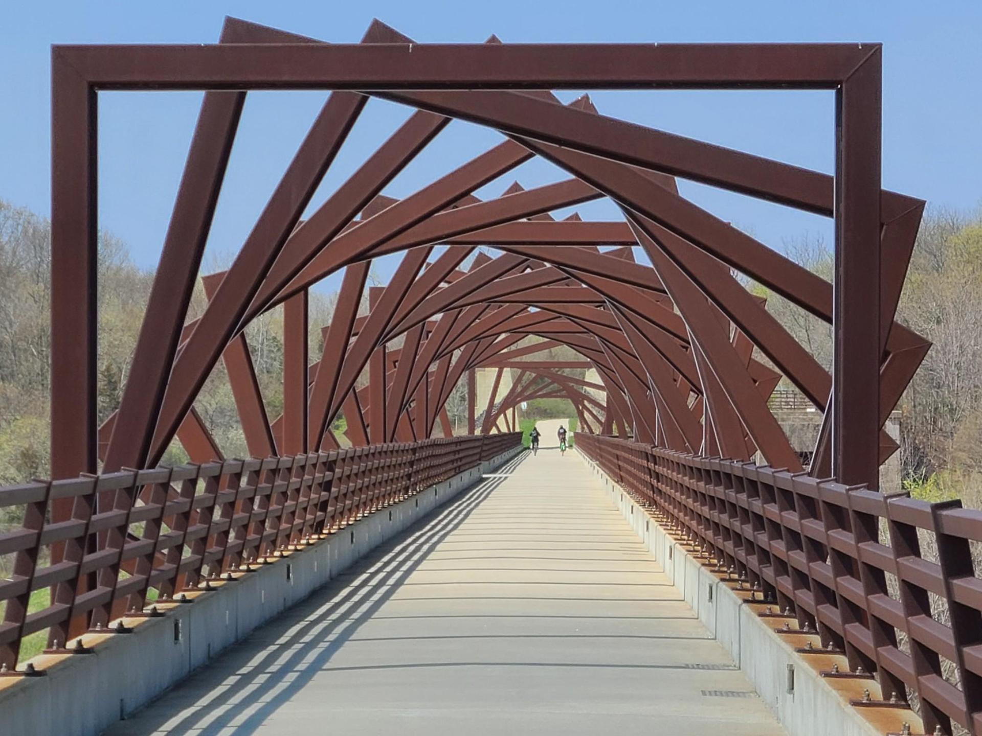Two people walk on a long, modern bridge with geometric rusty metal arches.