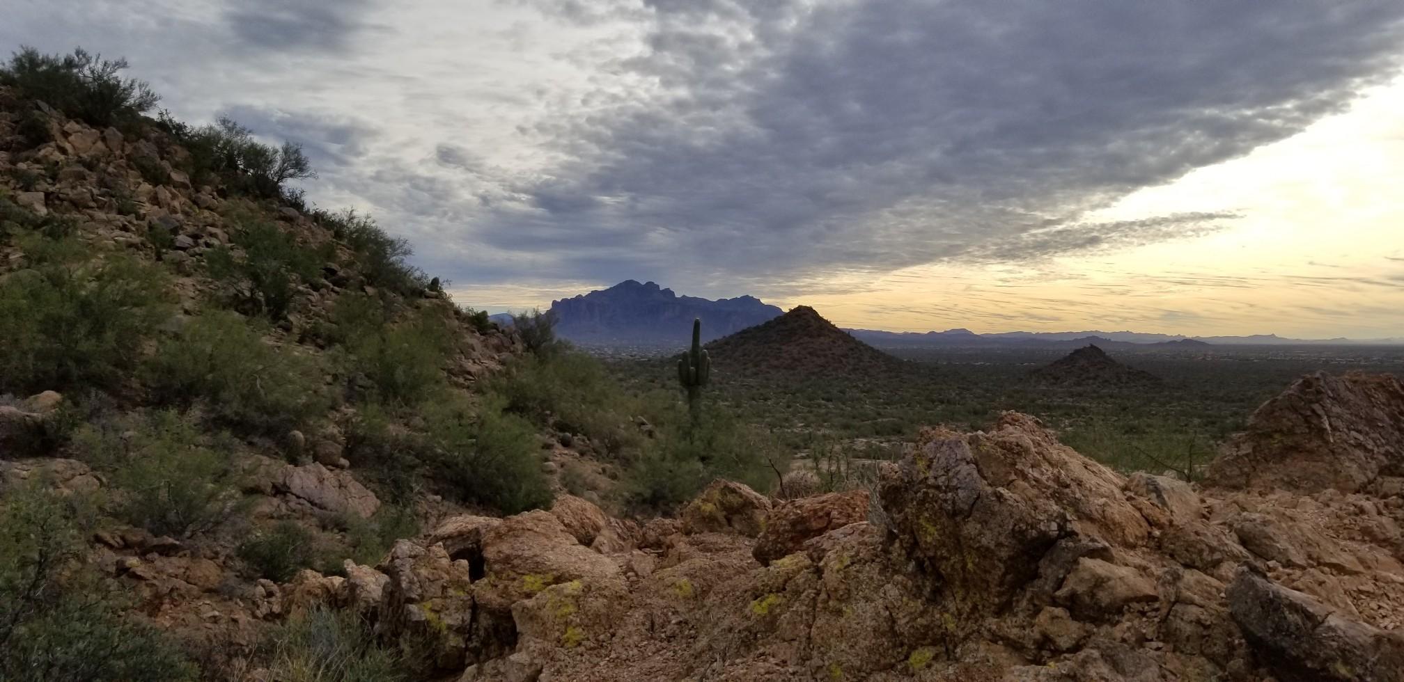 Vast desert panorama with rocky foreground, distant mountains, and dramatic cloudy sky.