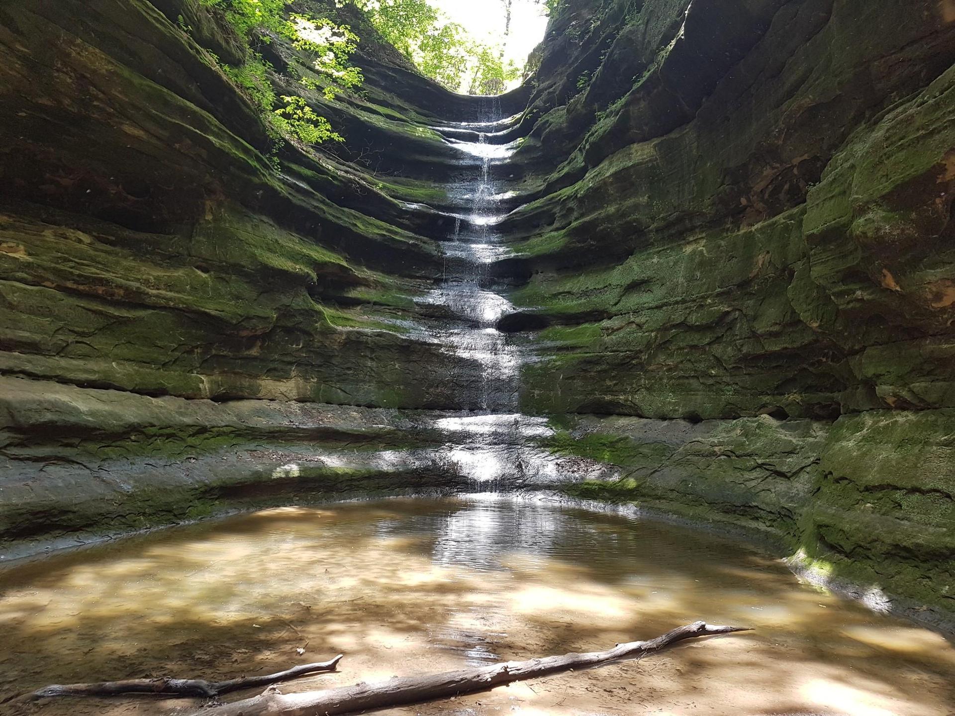 Tiered waterfall cascades into a shallow pool within a mossy rock canyon.