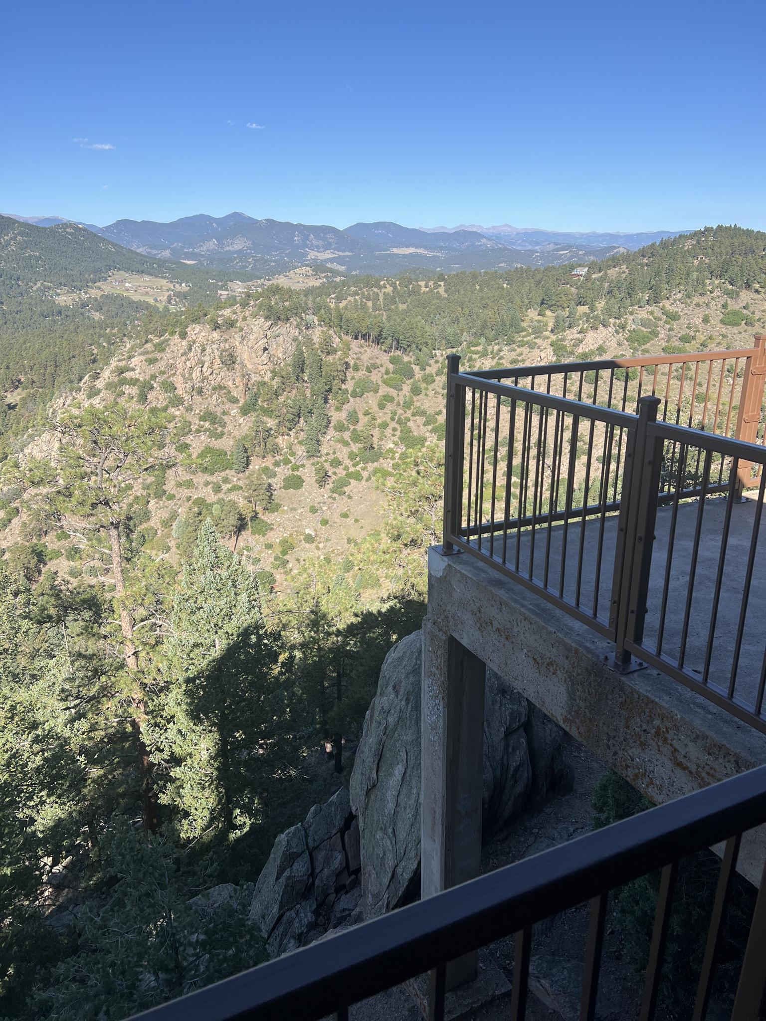 Observation deck railing overlooking a deep, sunny valley with forested mountains.