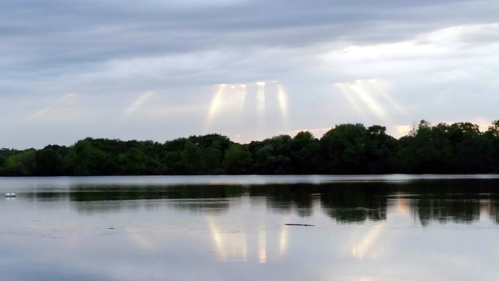 Sun rays pierce through clouds over a calm lake and forested shore.