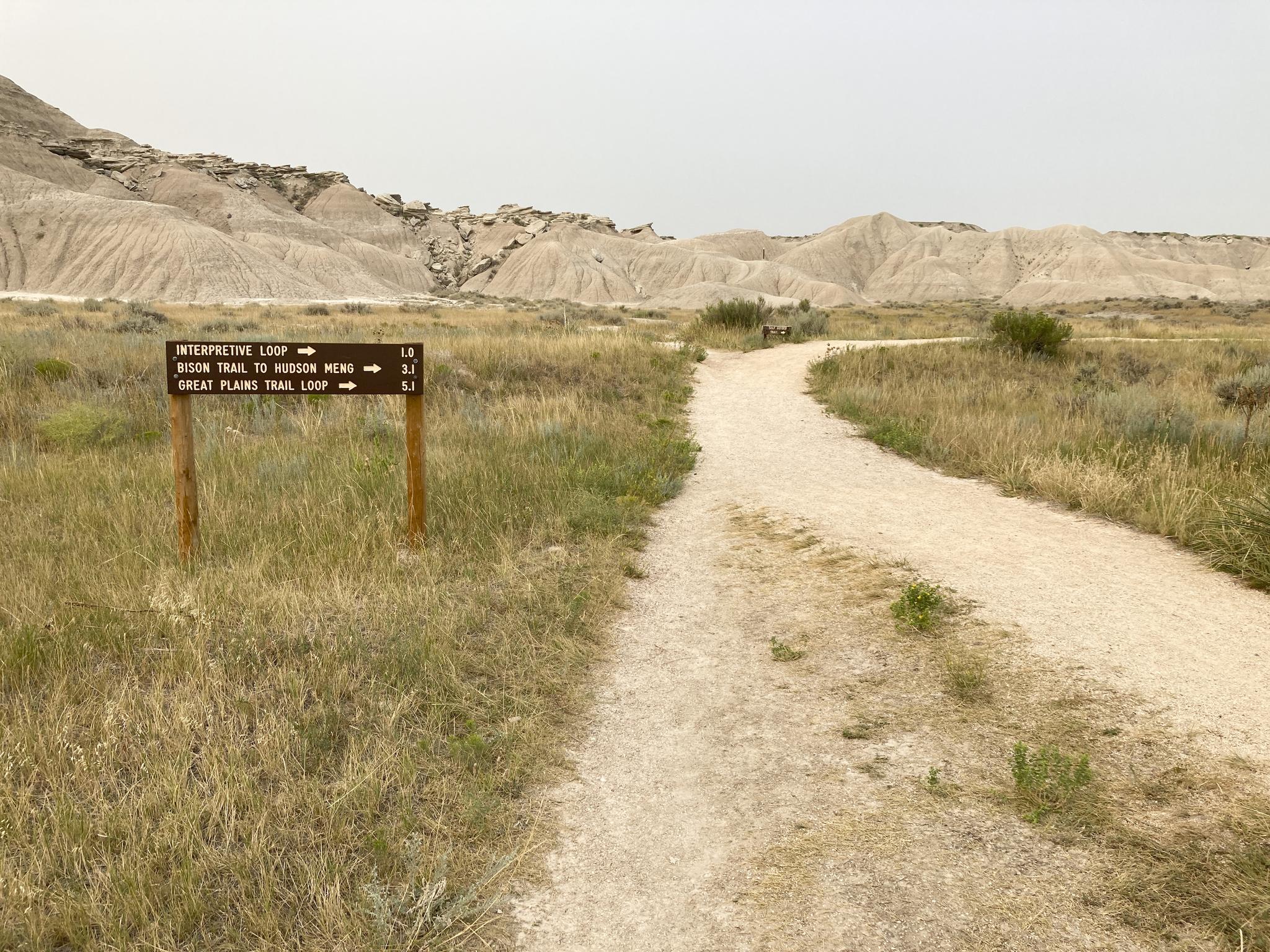 A dirt path and trail sign in dry grass with eroded hills under a hazy sky.