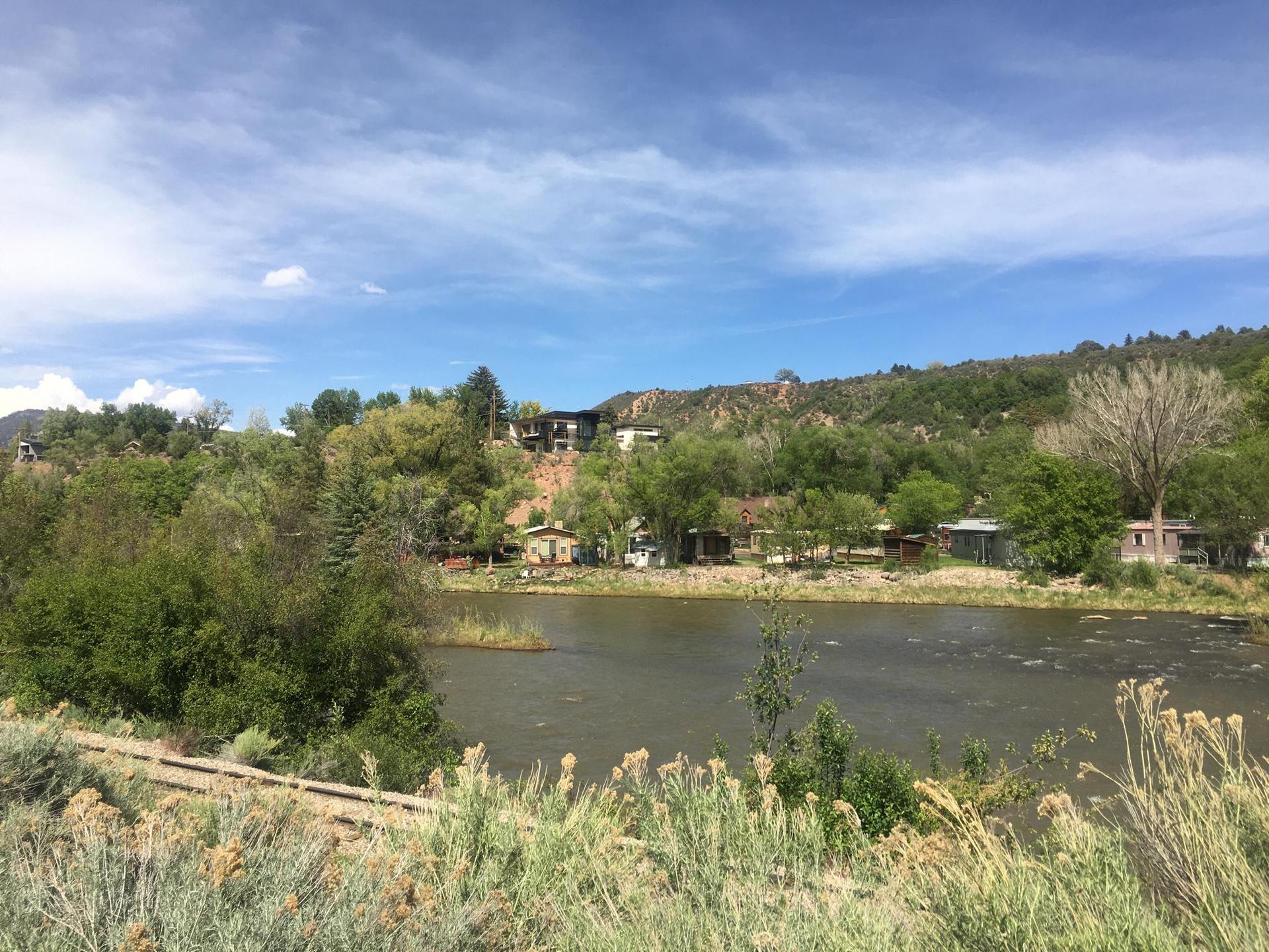A river flows past houses on a green hillside under a partly cloudy sky.
