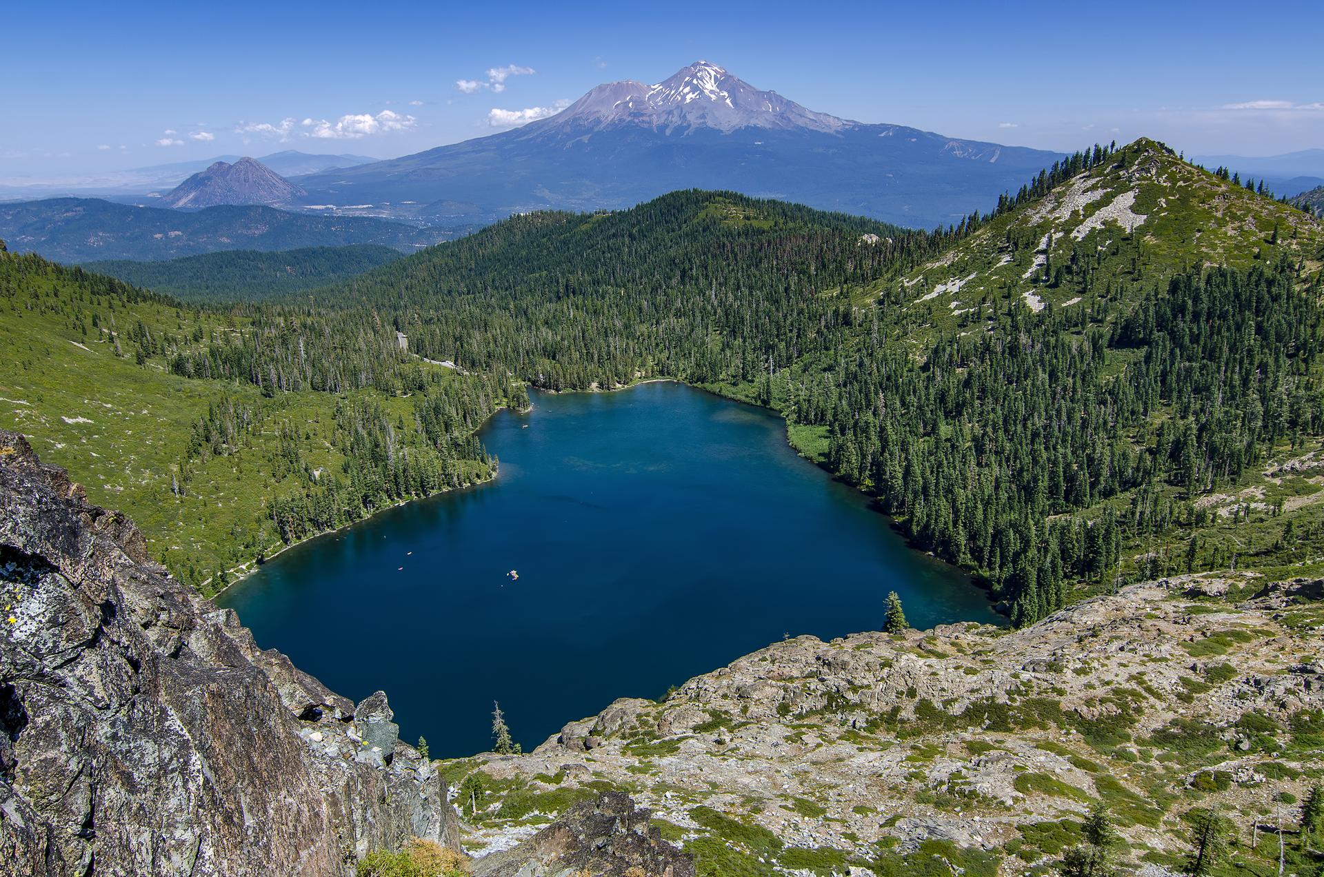 Deep blue mountain lake surrounded by green forest, with a distant snow-capped peak.
