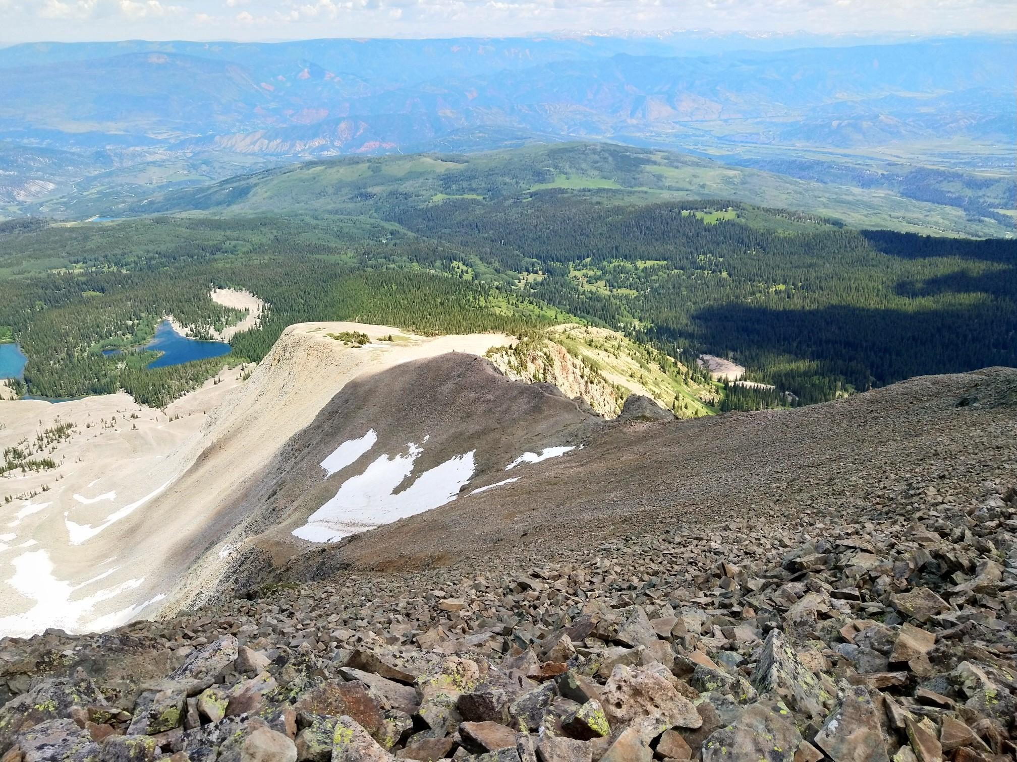 Steep rocky mountain ridge with snow patches, distant forests, and blue lakes.