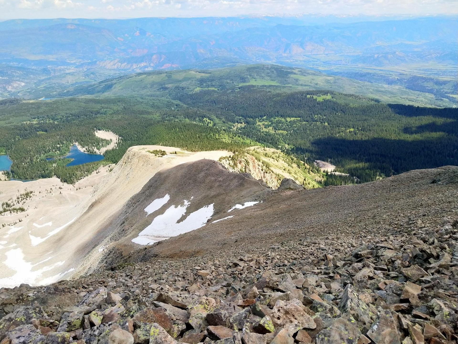 Steep rocky mountain ridge with snow patches, distant forests, and blue lakes.