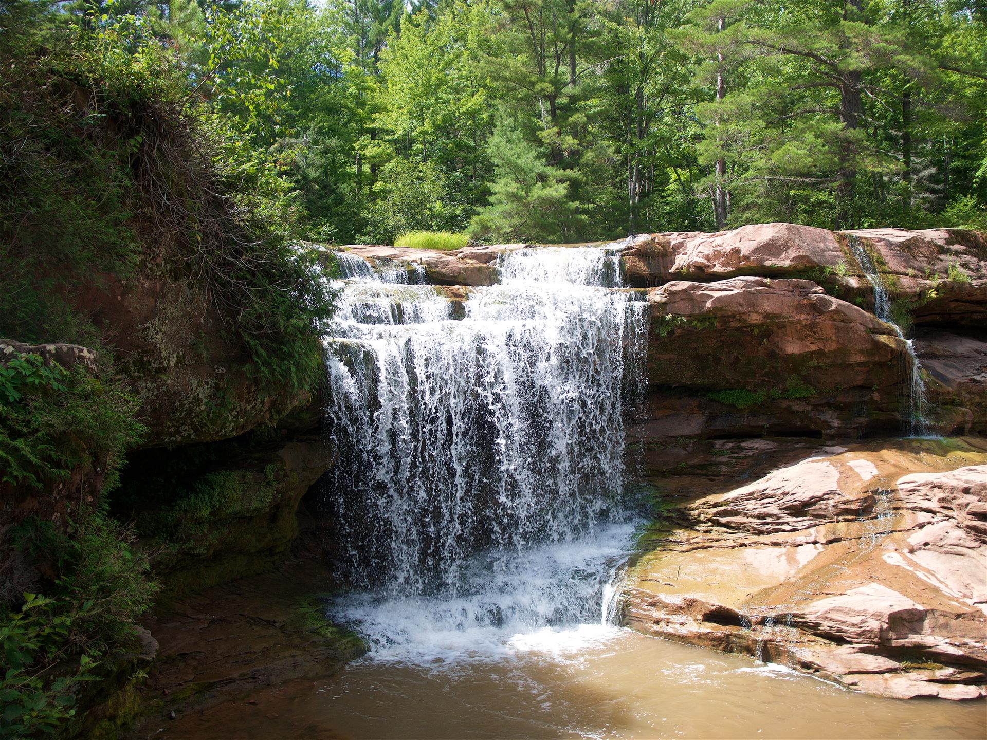 Waterfall cascades over red rocks into a pool, surrounded by green trees.