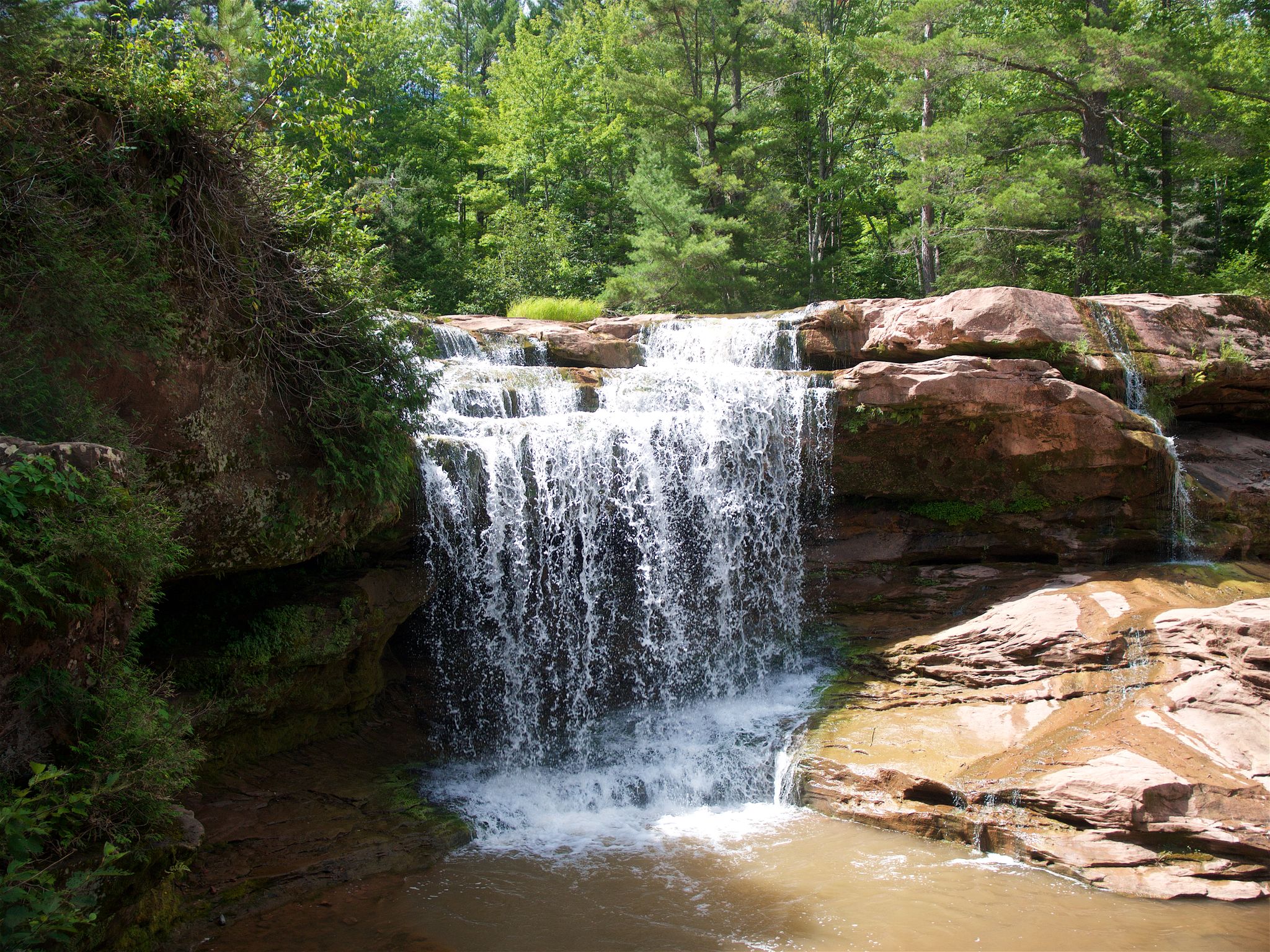 Waterfall cascades over red rocks into a pool, surrounded by green trees. - O Kun de Kun Falls via North Country Trail