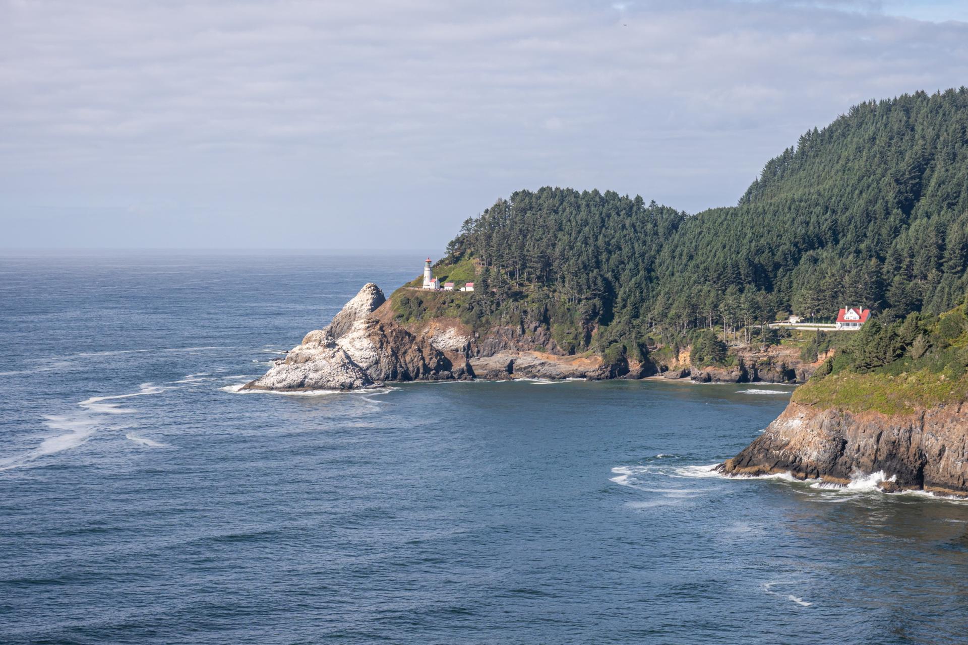Blue ocean and forested cliffs with a lighthouse on a rocky headland.