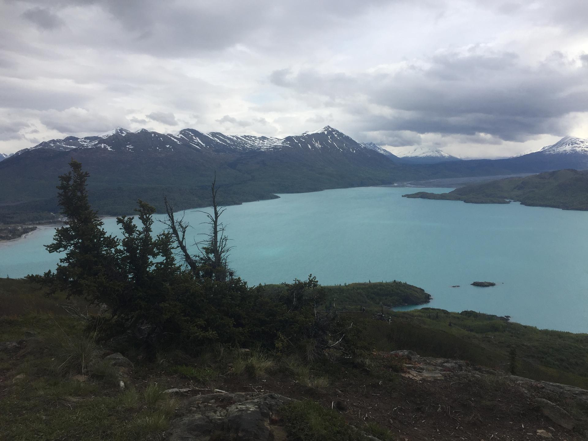 Overcast view of a turquoise lake surrounded by snow-capped mountains.