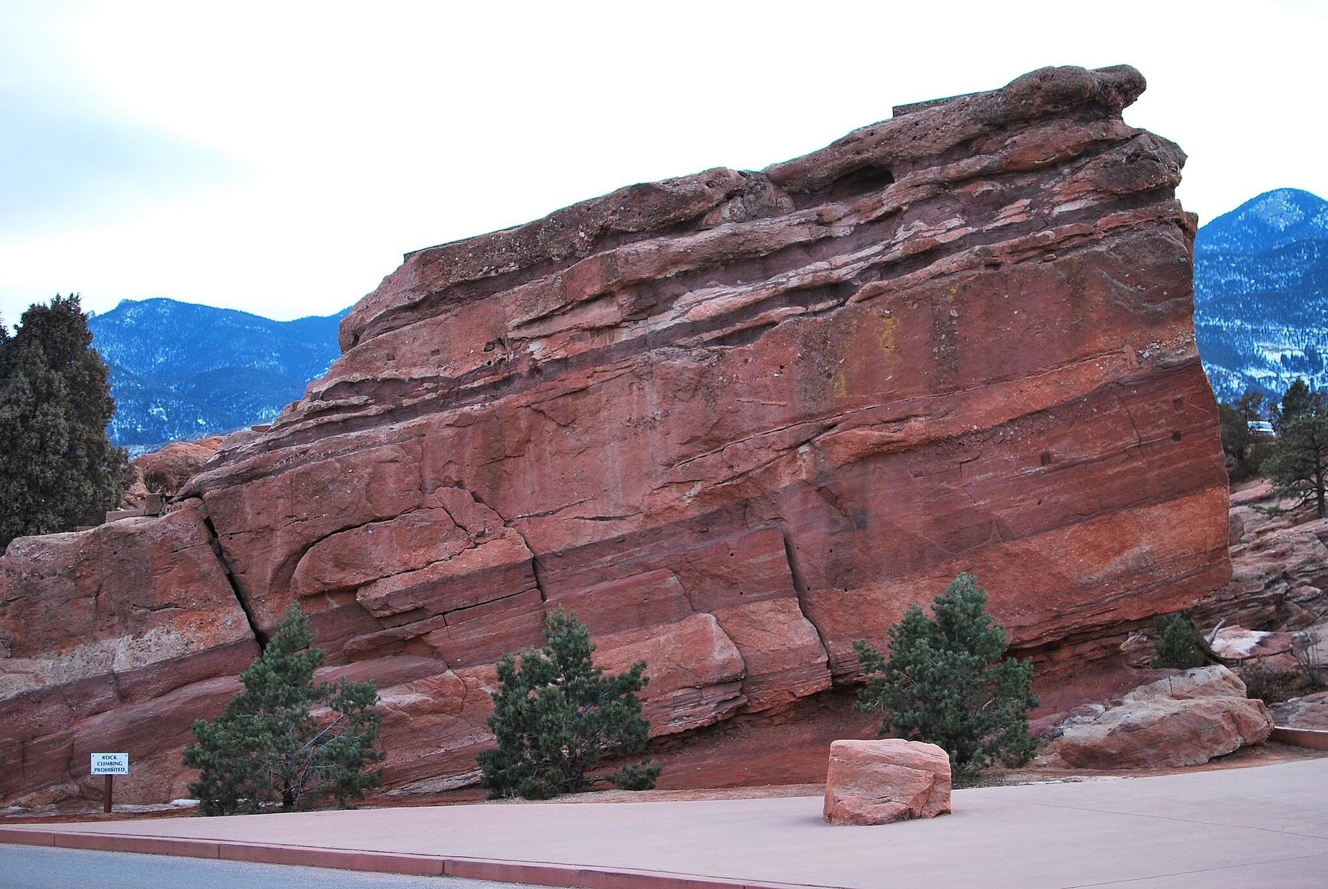 Enormous layered red rock formation with small trees under an overcast sky.