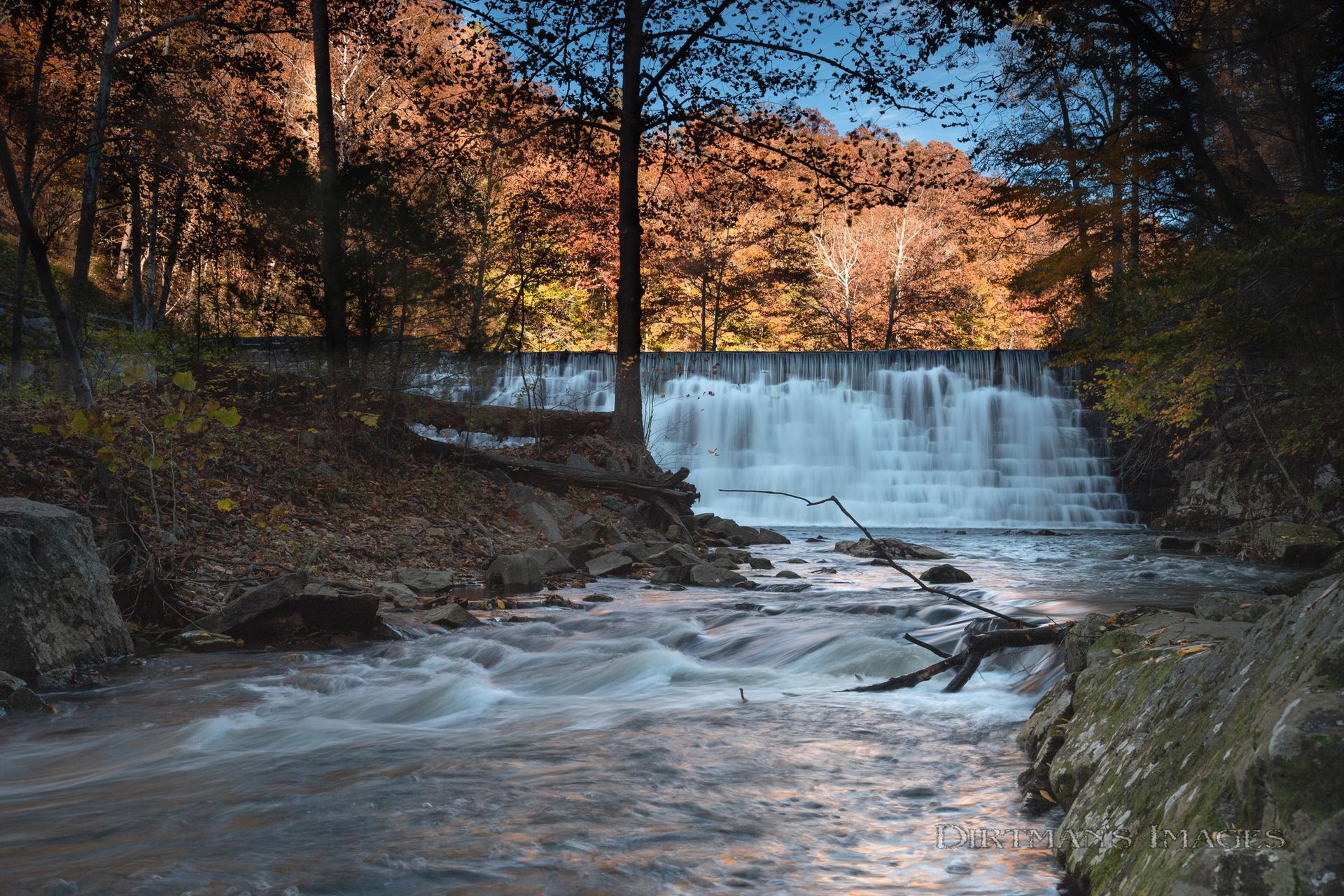 River flowing past a waterfall, surrounded by vibrant autumn trees at dusk.