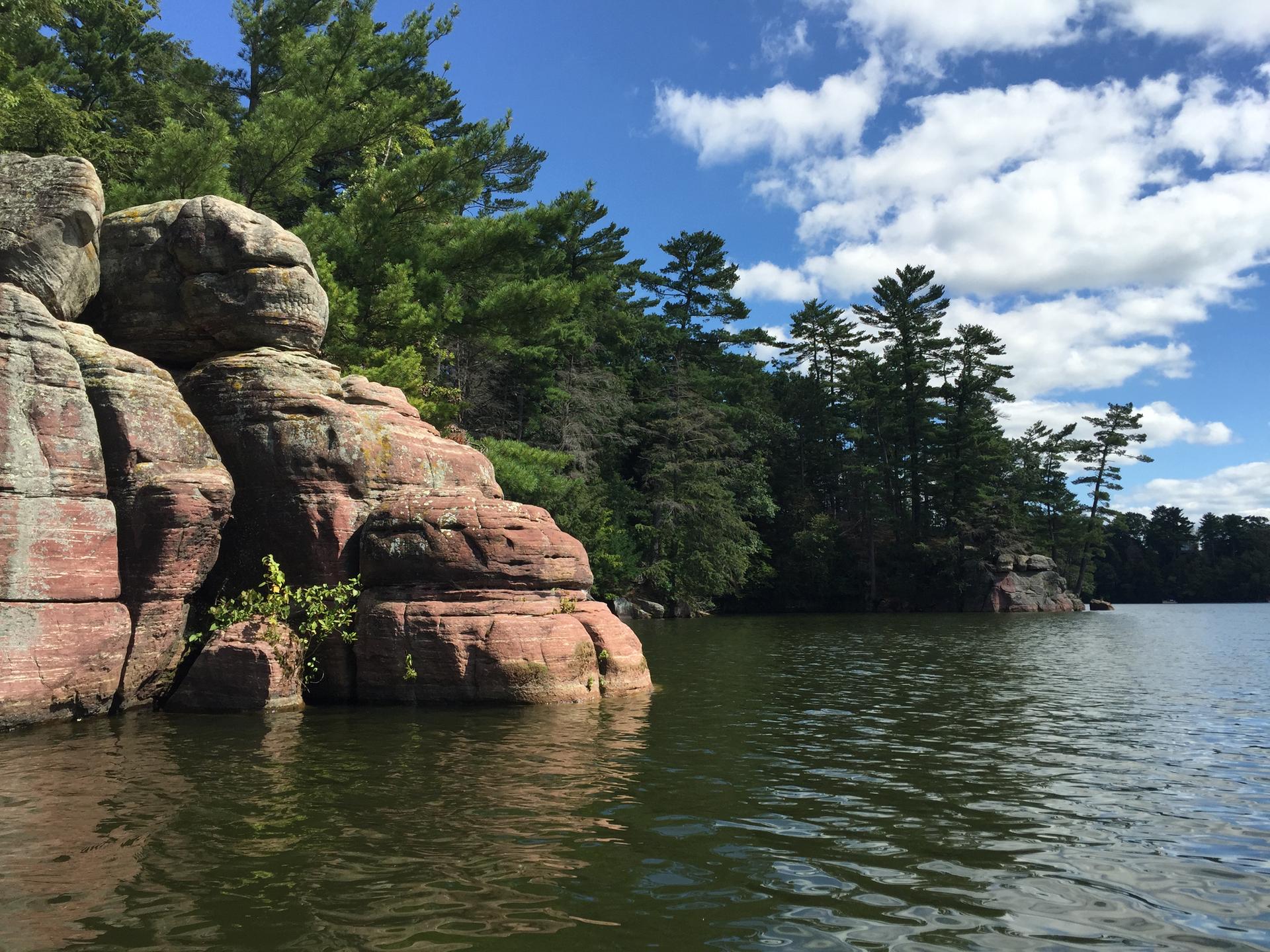 Red rock formations rise from a calm lake, surrounded by green pine trees.