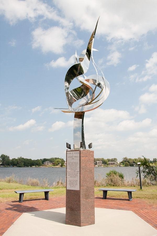 A tall, abstract metal sculpture stands by a lake under a bright sky.