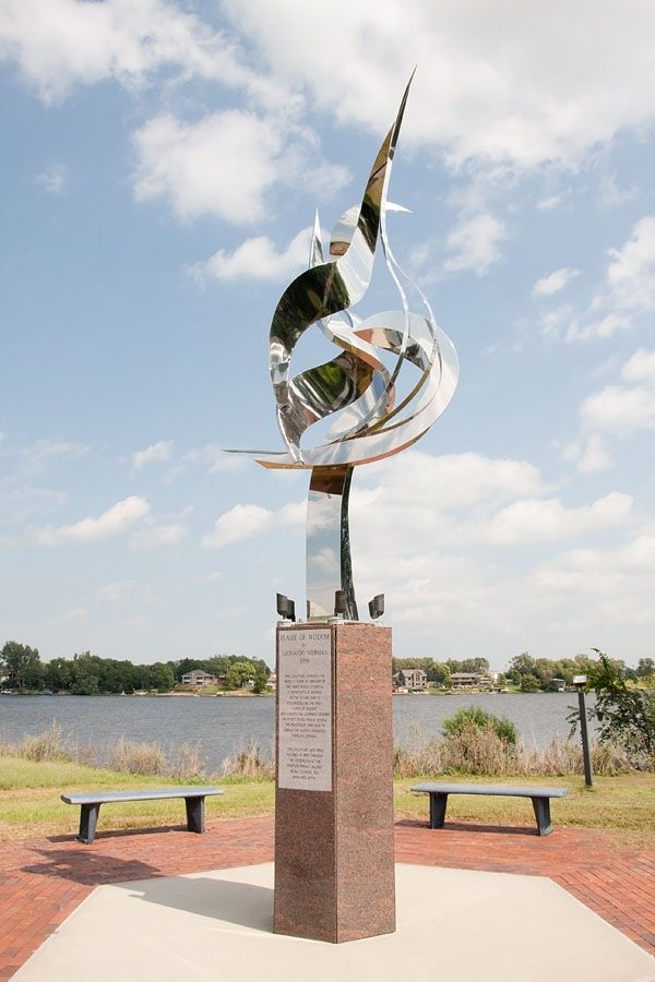 A tall, abstract metal sculpture stands by a lake under a bright sky. - Lake Mitchell – Lake Mitchell loop from Mitchell Township