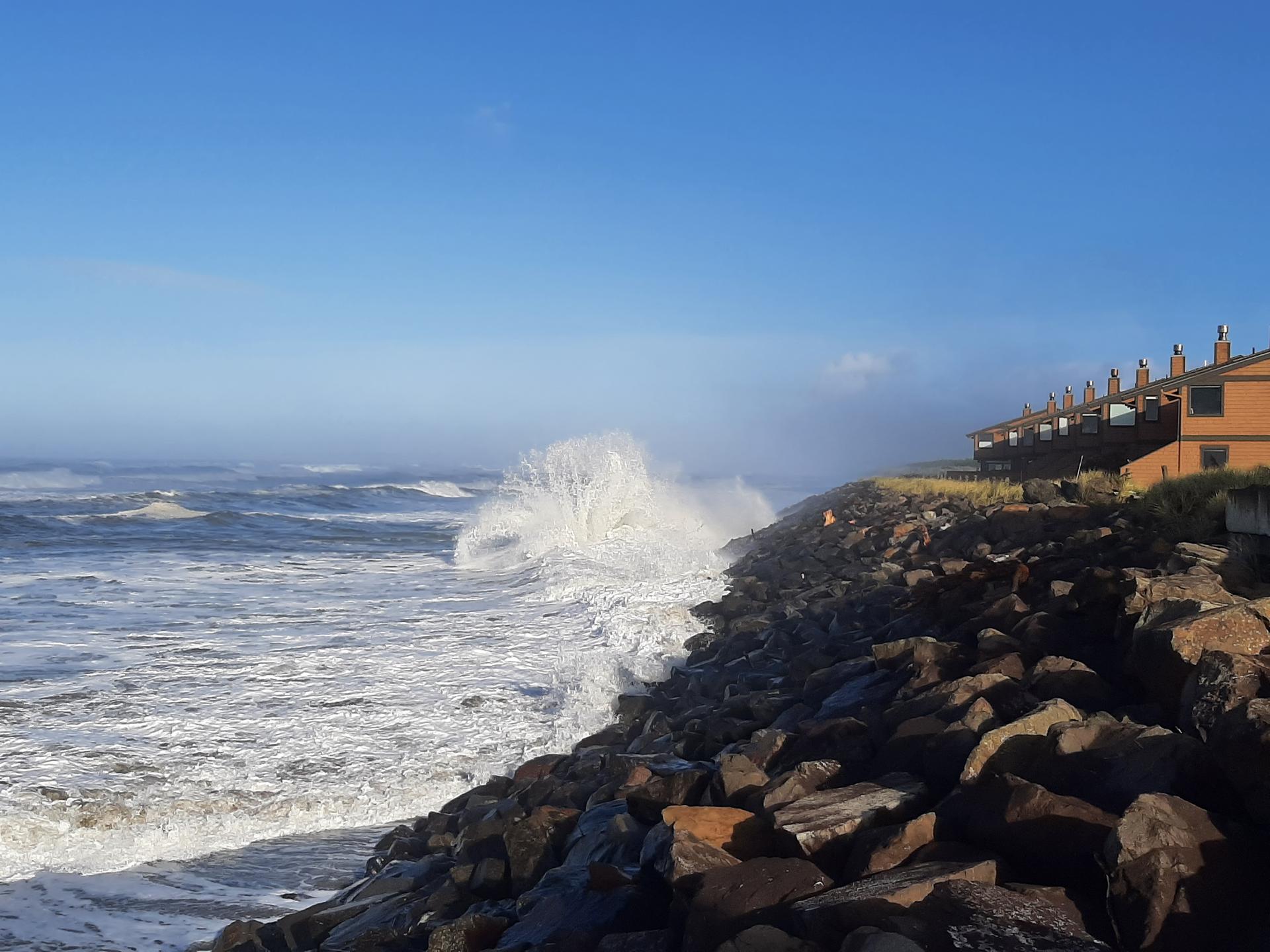 Huge ocean wave crashing against a rocky shore under a clear blue sky.