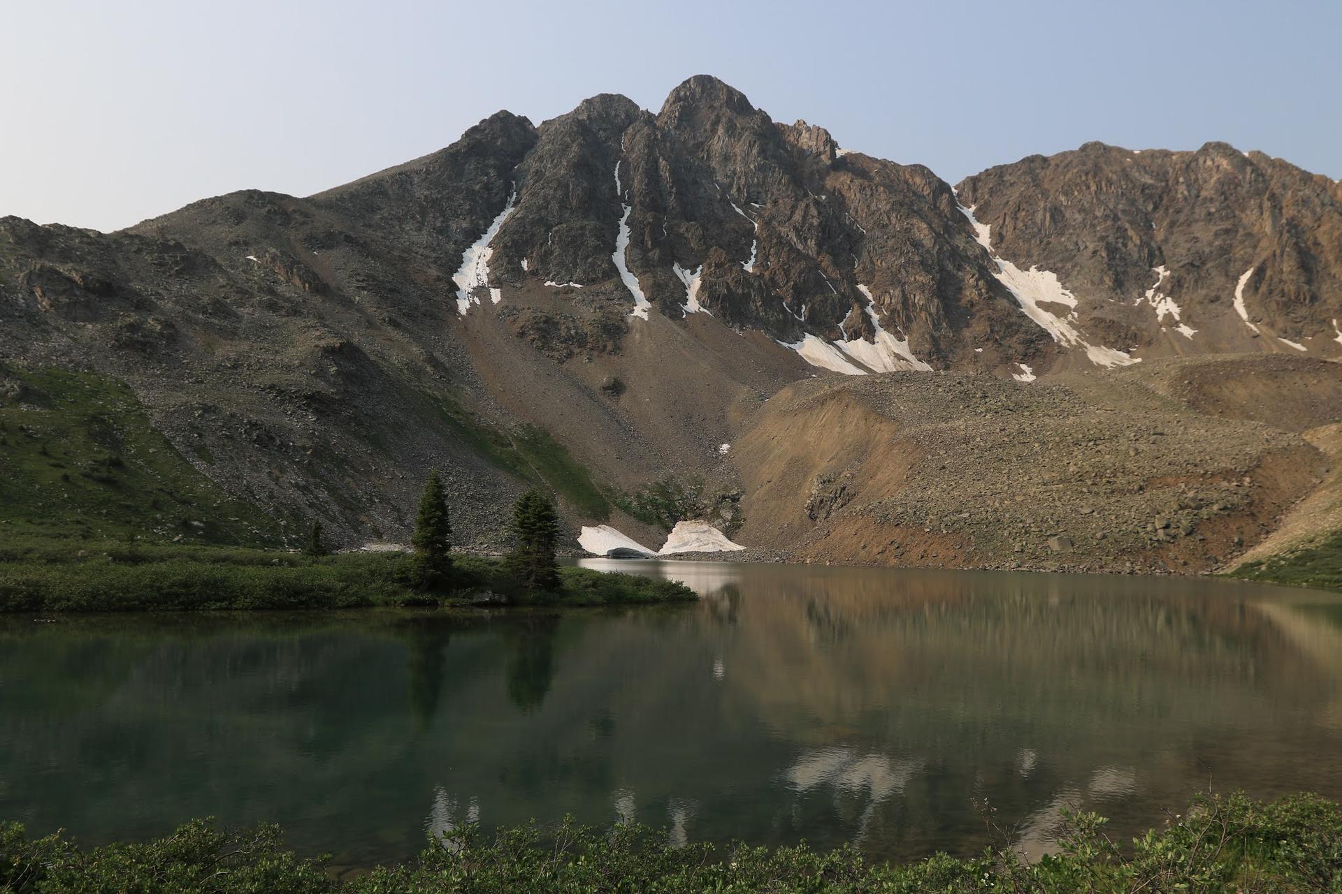 Calm mountain lake reflecting rocky peaks with snow patches under soft light.