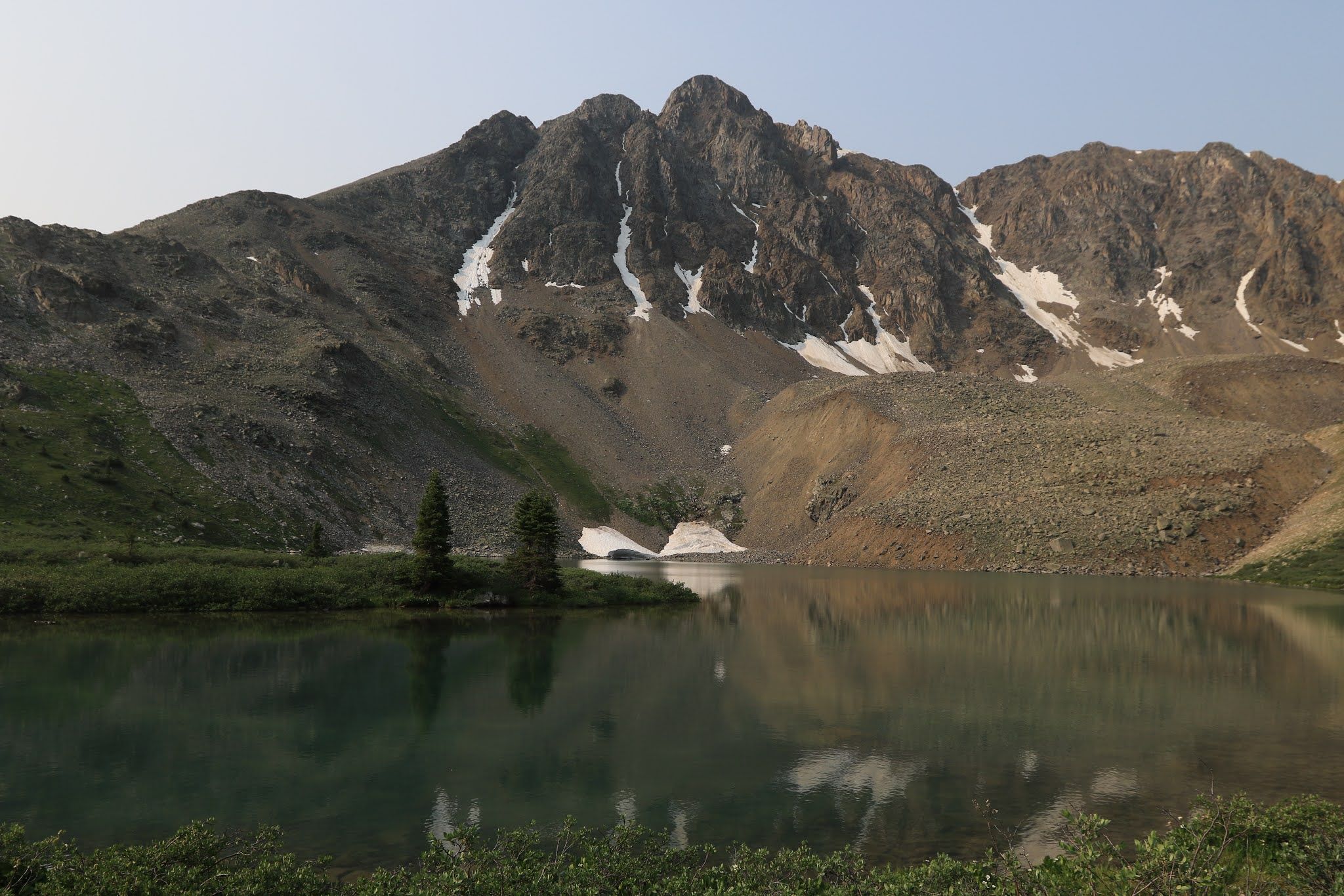 Calm mountain lake reflecting rocky peaks with snow patches under soft light. - Lincoln Creek to Petroleum and Anderson Lakes Trail