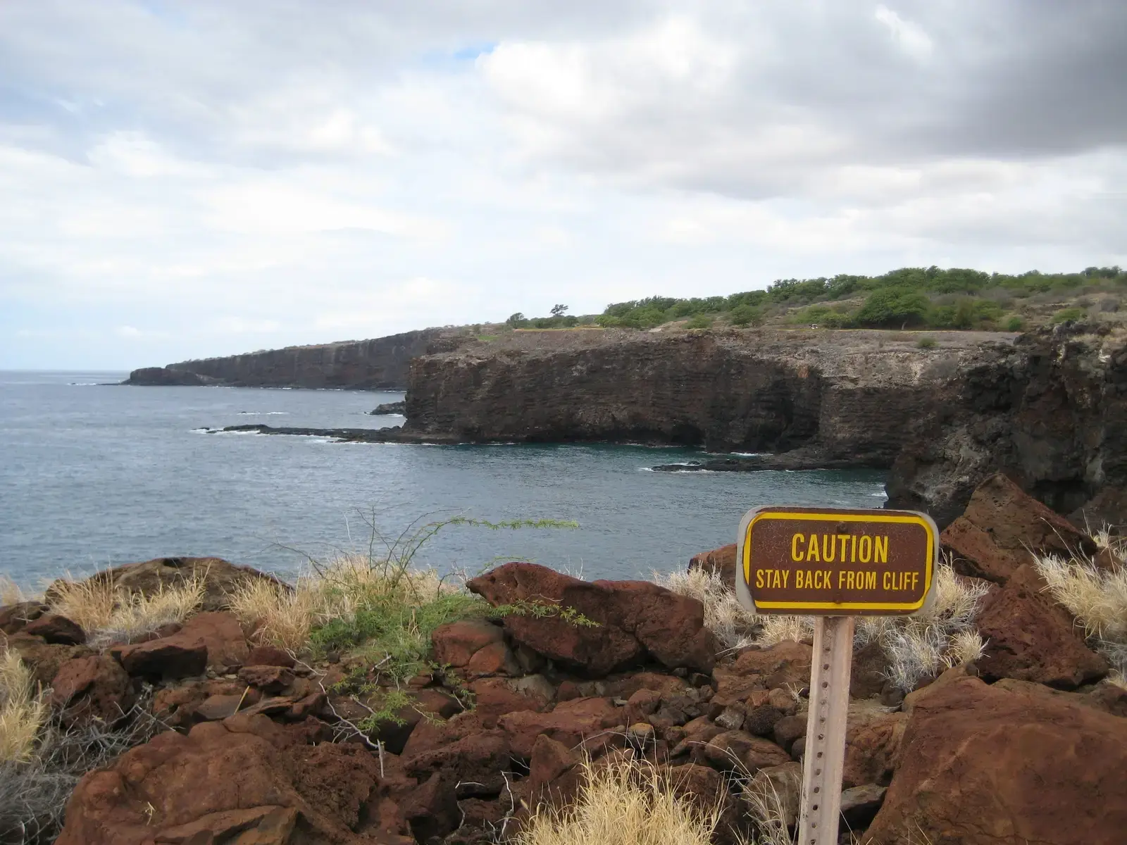 Caution sign on rocky ground near rugged coastal cliffs and calm ocean.